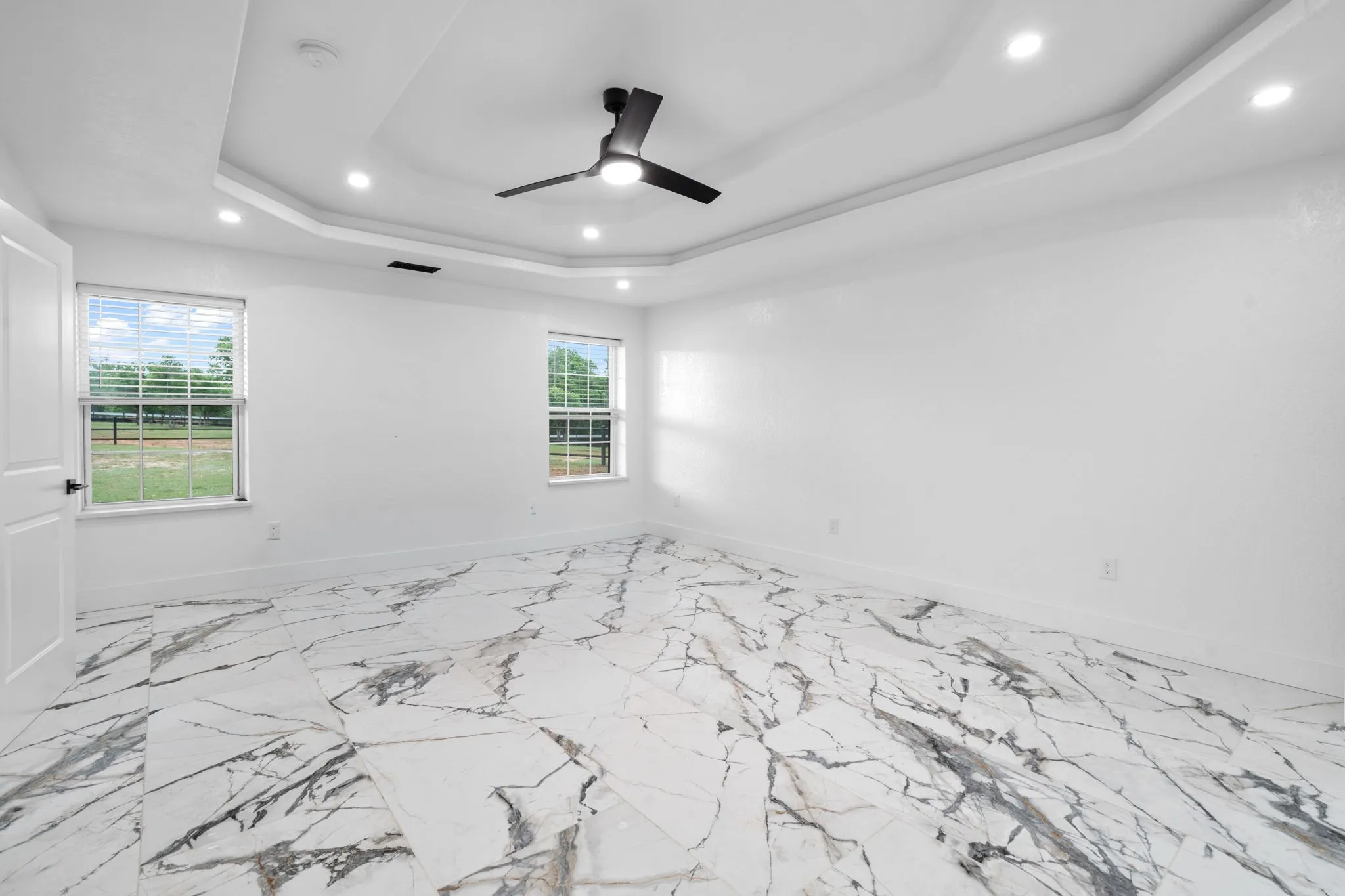 Spacious Primary bedroom featuring large format tile flooring with a marble-effect pattern, a tray ceiling with recessed lighting, and a modern ceiling fan