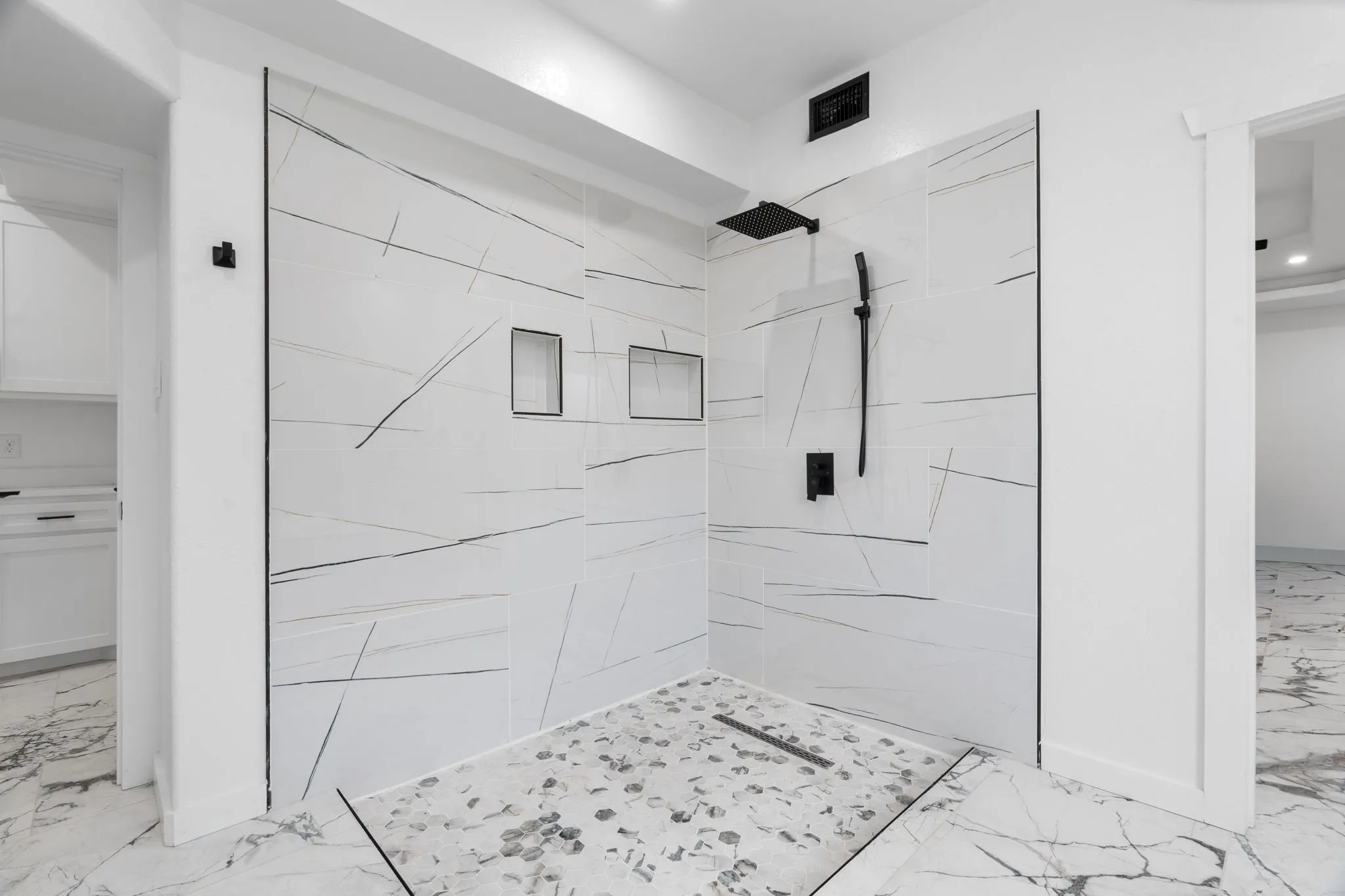 Walk-in shower featuring large format white tile with linear veining, pebble mosaic floor, dual recessed niches, matte black rainfall showerhead, and handheld shower sprayer