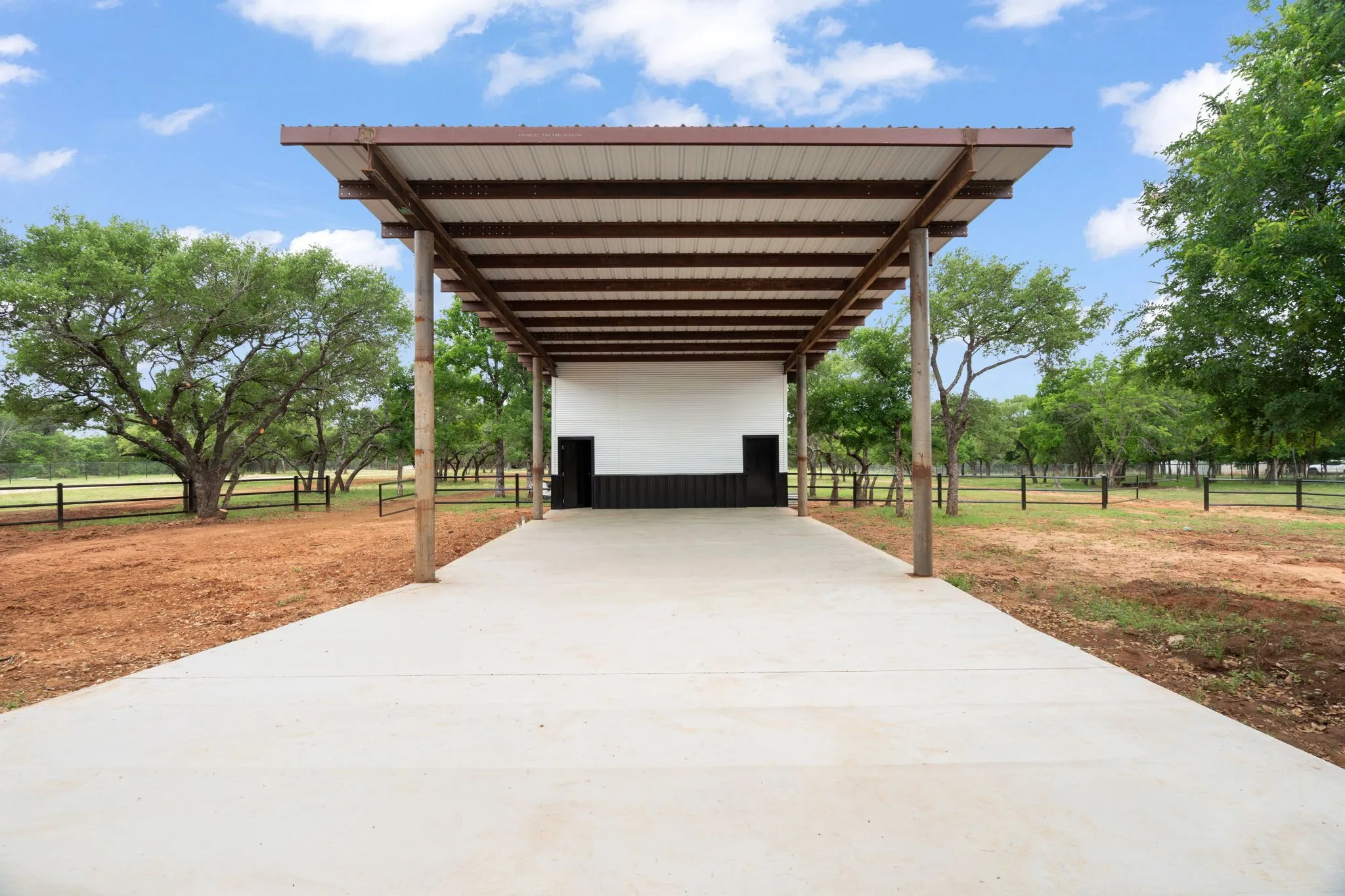 Covered concrete pad featuring a metal roof structure supported by wood posts, a solid white wall with a black base, and surrounding mature trees