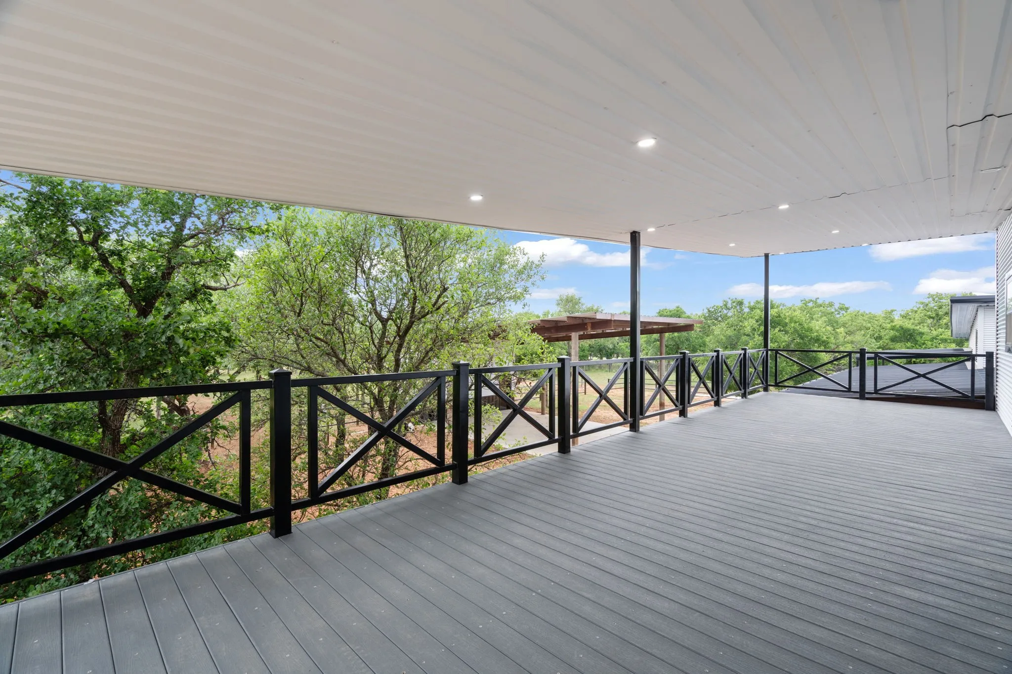 Expansive covered deck featuring composite decking, recessed lighting, black X-pattern railing, and a corrugated​​‌​​​​‌​​‌‌​‌‌​​​‌‌​‌​‌​‌​​​‌​​ ceiling