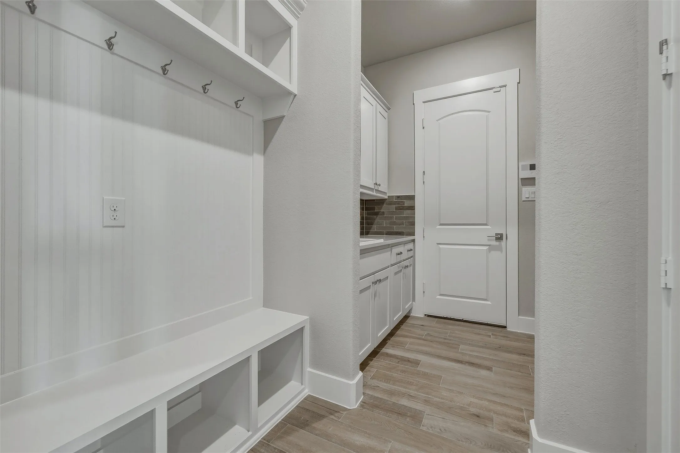 Built-in mudroom bench with storage cubbies and coat hooks, wood-finish flooring, white cabinetry with a tile backsplash, and a paneled interior door
