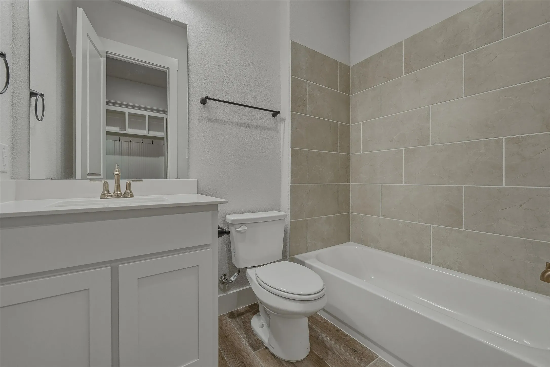 Vanity with white cabinetry and integrated sink, chrome faucet, bathtub with beige tile surround, and wood-finish flooring