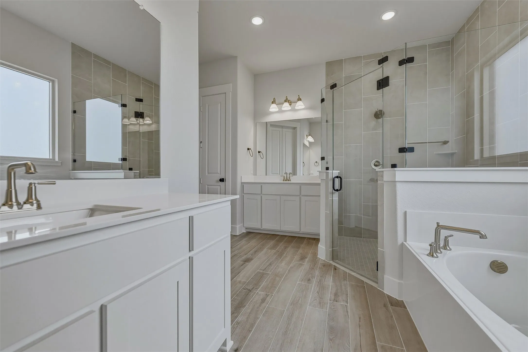 White cabinetry with integrated sinks, brushed nickel fixtures, a glass-enclosed shower, a built-in tub, and wood-finish tile flooring