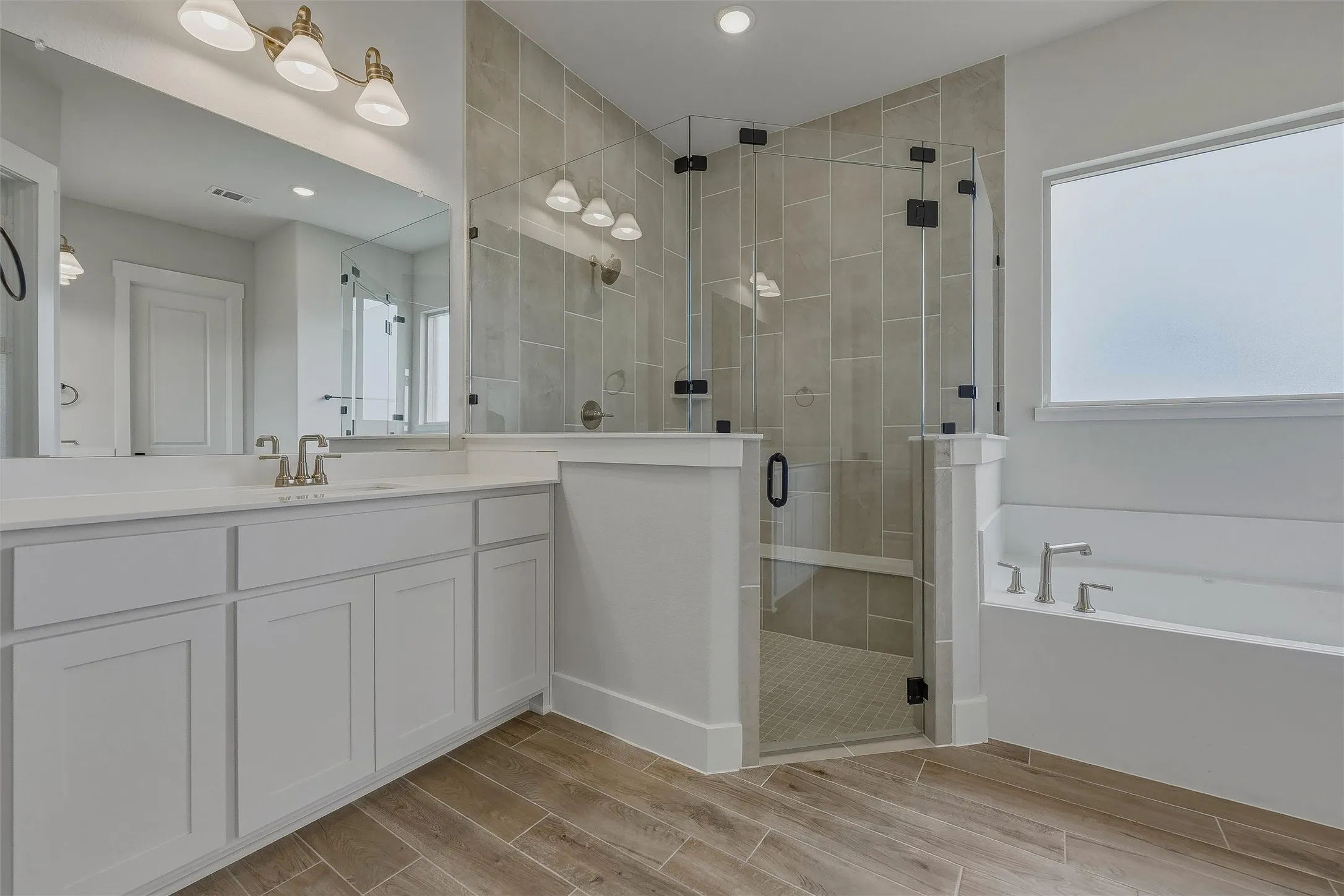 Dual vanity with white cabinetry, wood-finish flooring, tiled walk-in shower with glass enclosure, and built-in soaking tub