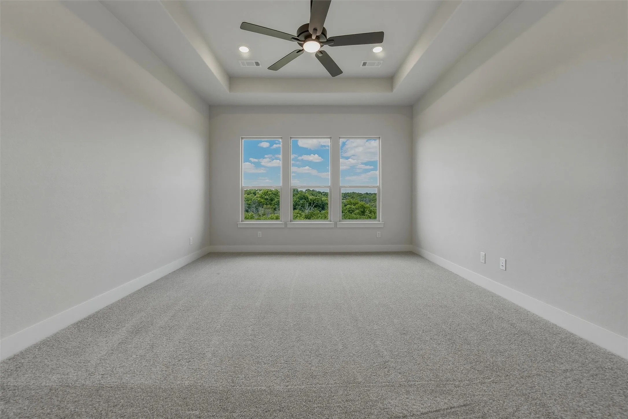 Spacious room featuring a tray ceiling with a ceiling fan, recessed lighting, carpeted flooring, and three windows offering expansive outdoor scenery