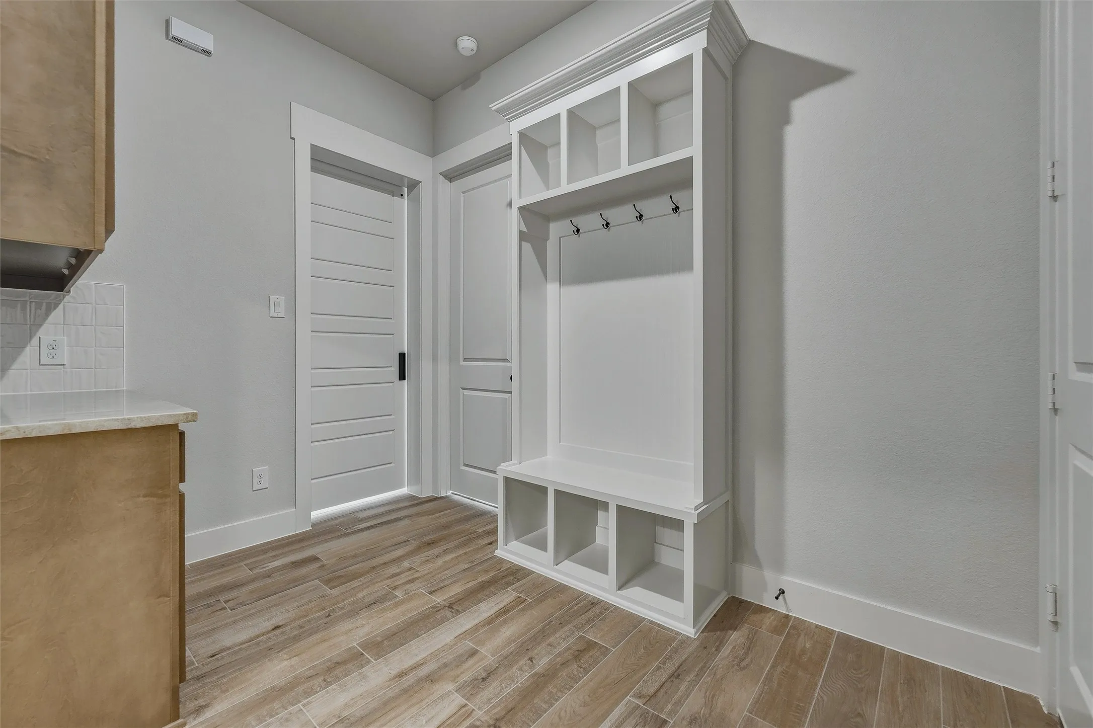 Wood-finish tile flooring, built-in white storage unit with hooks and cubbies, white shaker-style doors, light wood-finish cabinetry, and white subway tile backsplash