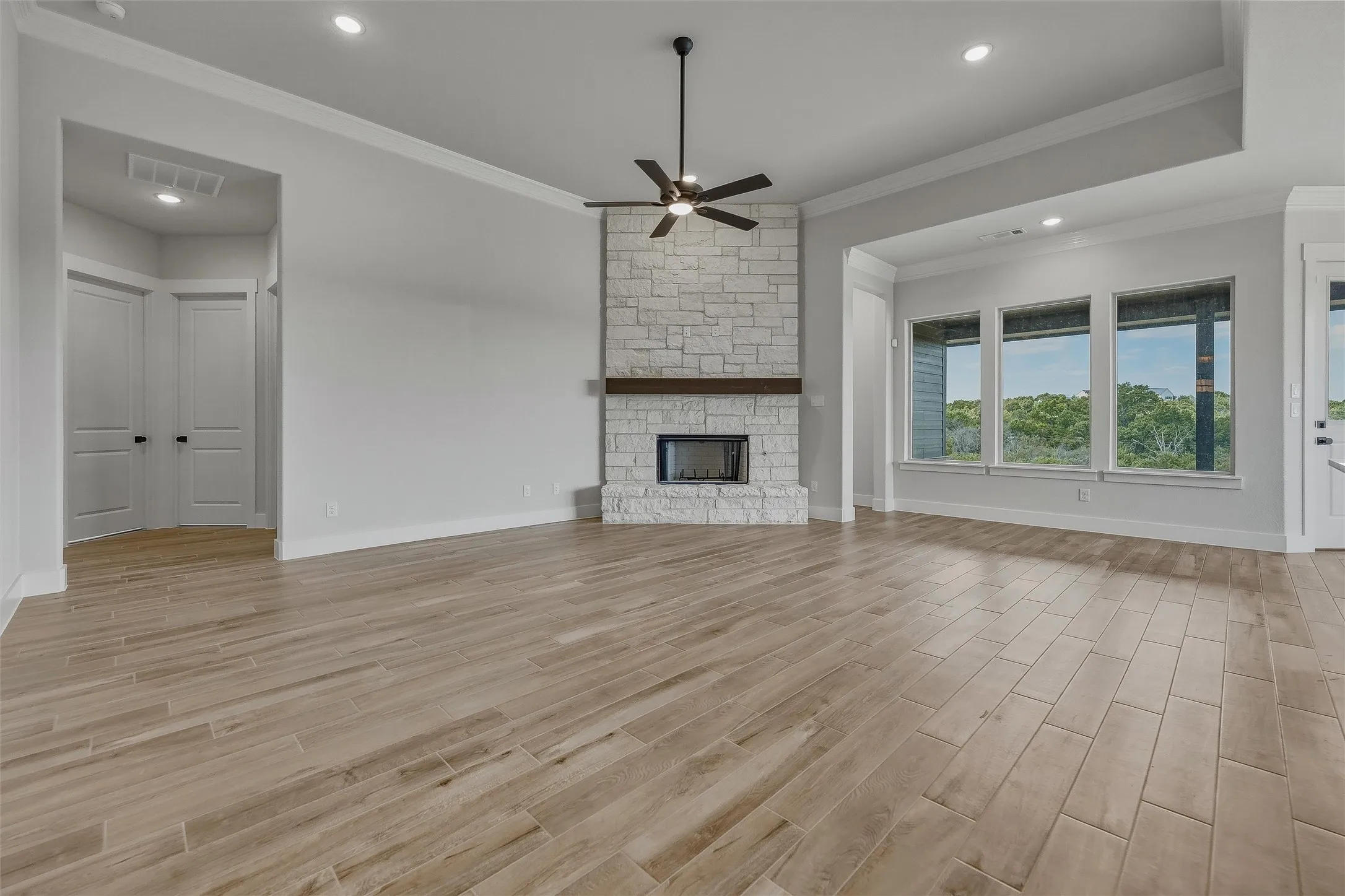 Spacious area featuring wood-finish tile flooring, a stone-faced fireplace with a wood mantel, a ceiling fan, recessed lighting, and multiple windows
