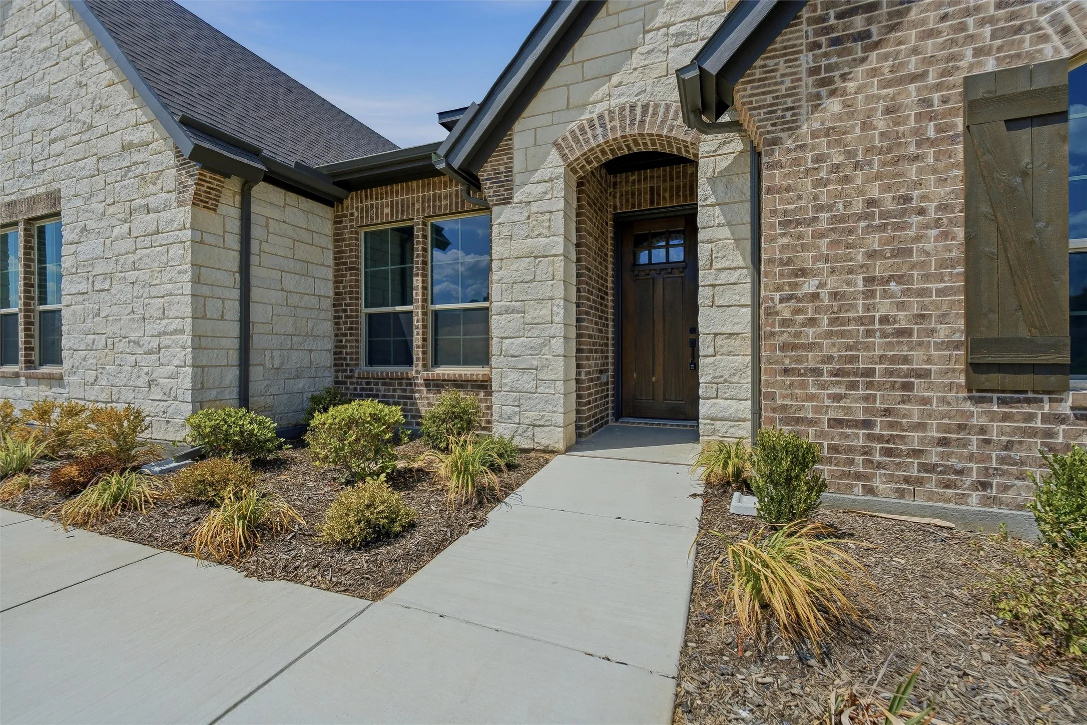 Architectural facade featuring a blend of light-colored stone and dark brick, a dark wood entry door with glass panels, and a concrete pathway