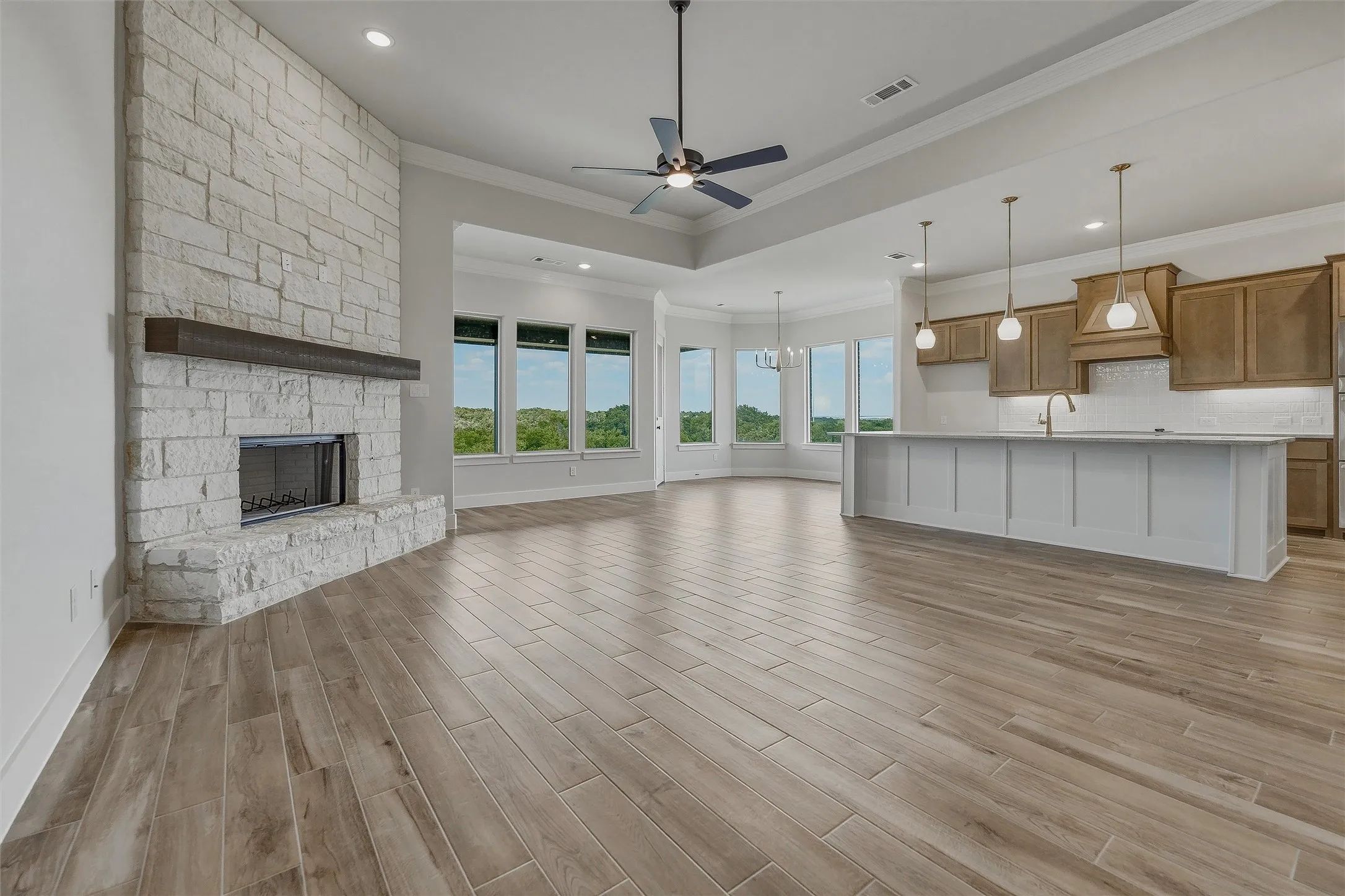 Expansive open-concept living area featuring a stone fireplace with a wood mantel, wood-finish flooring, and a kitchen island with pendant lighting