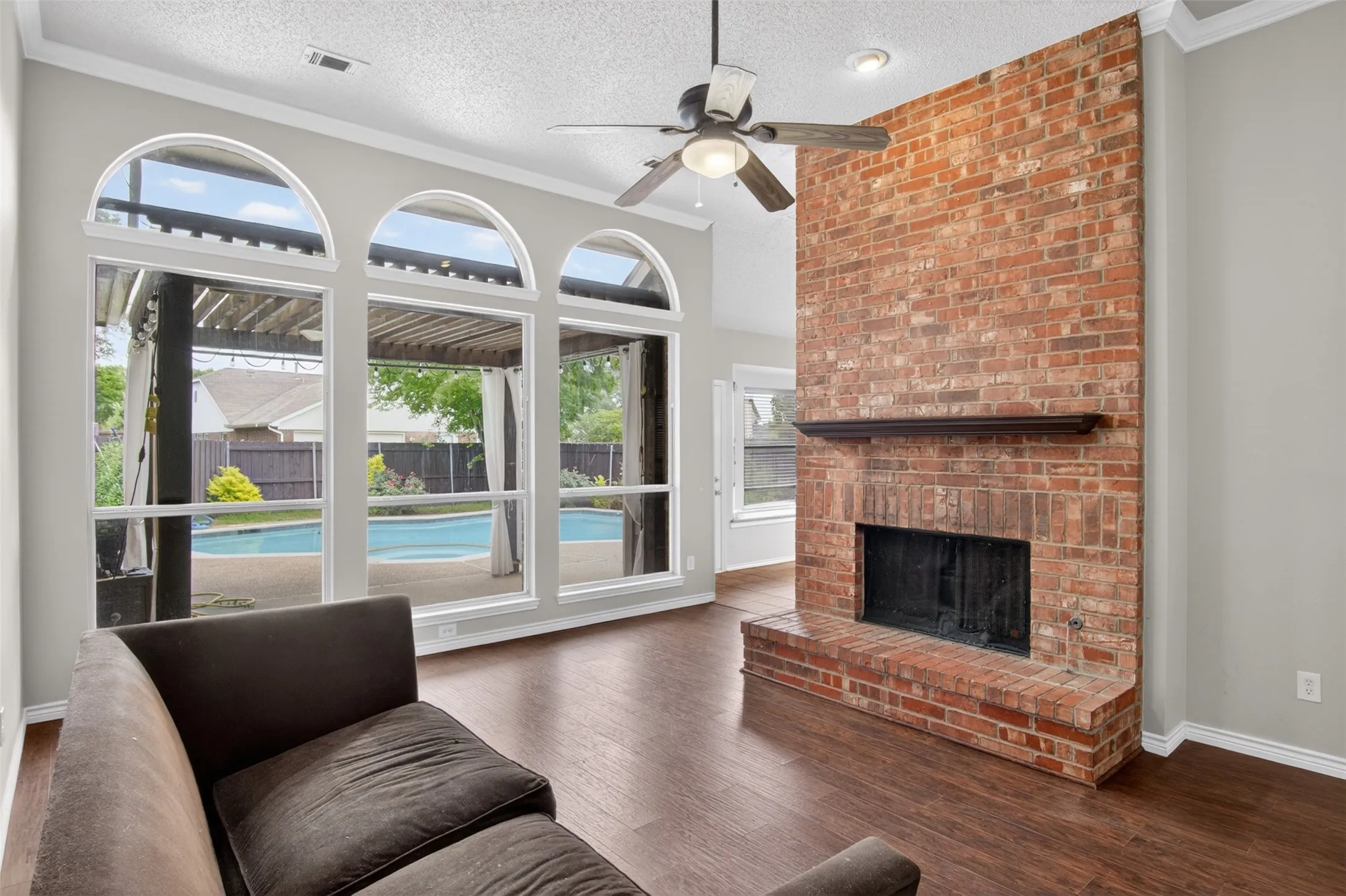 Living area featuring dark wood floors, a prominent brick fireplace with a mantel, and arched windows that offer views of the outdoor pool area