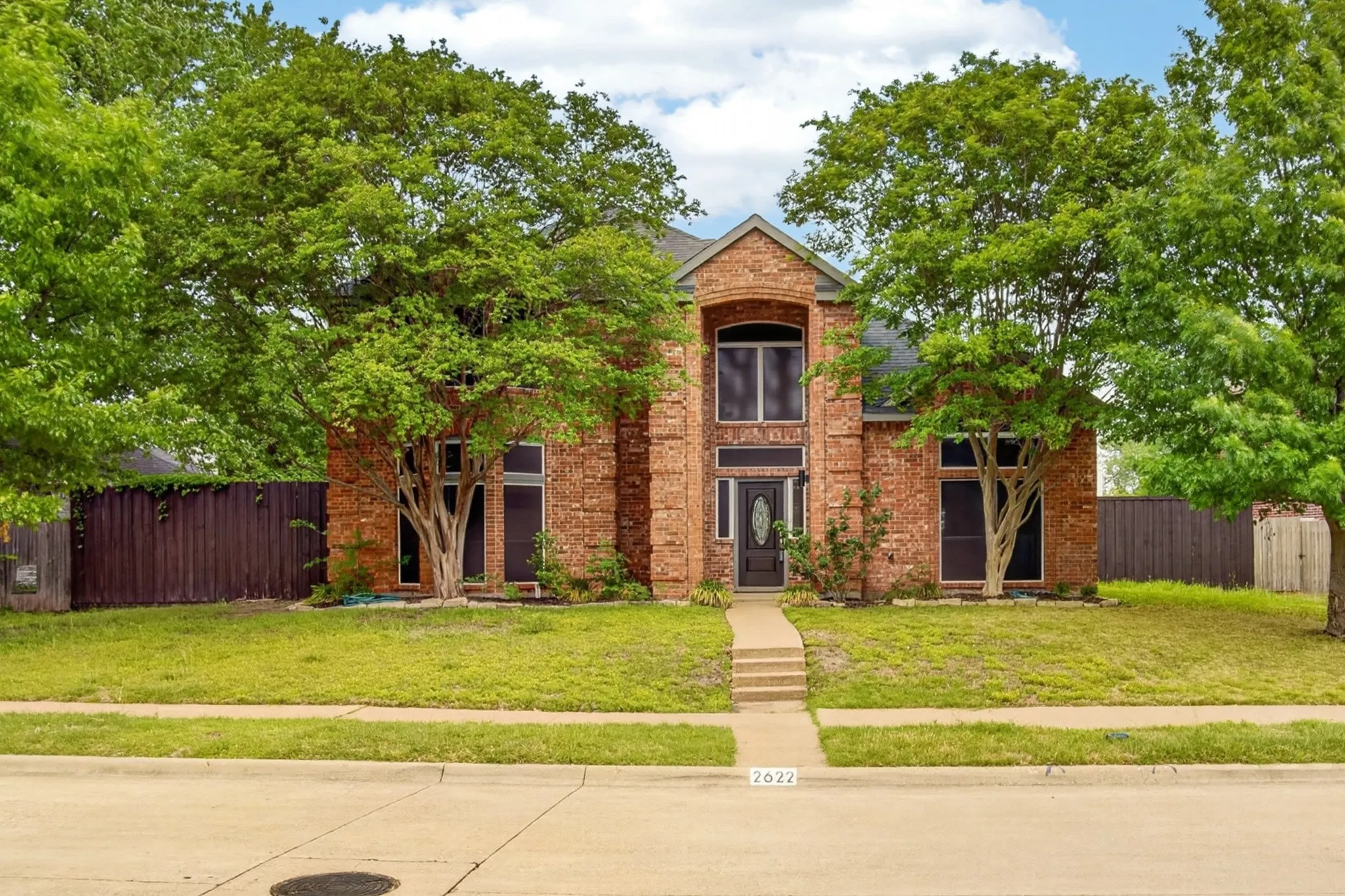 The property features a brick exterior, a well-maintained lawn, and mature trees in the front yard