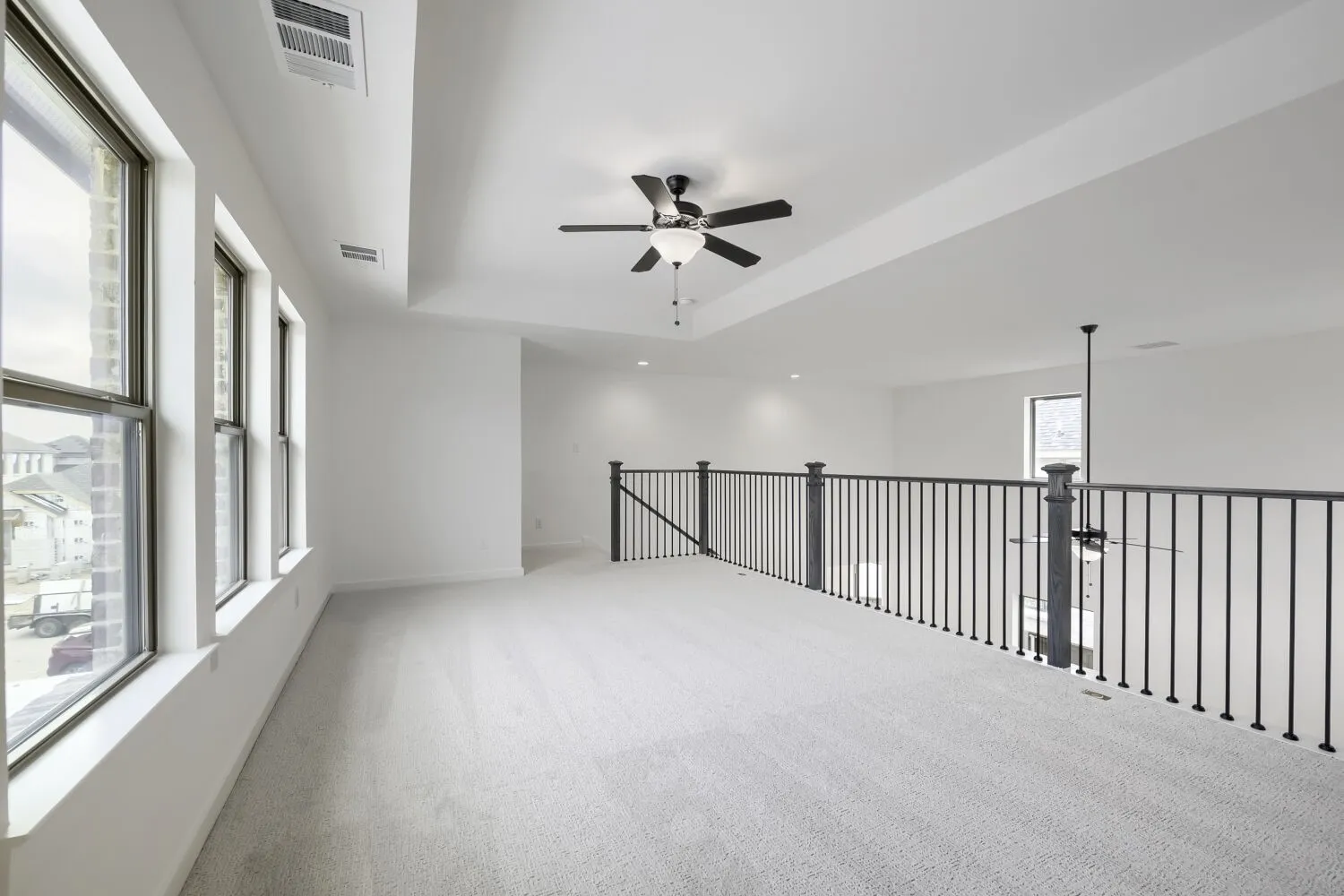 Carpeted loft space featuring a black iron spindle railing, multiple windows with white trim, a ceiling fan, and recessed lighting