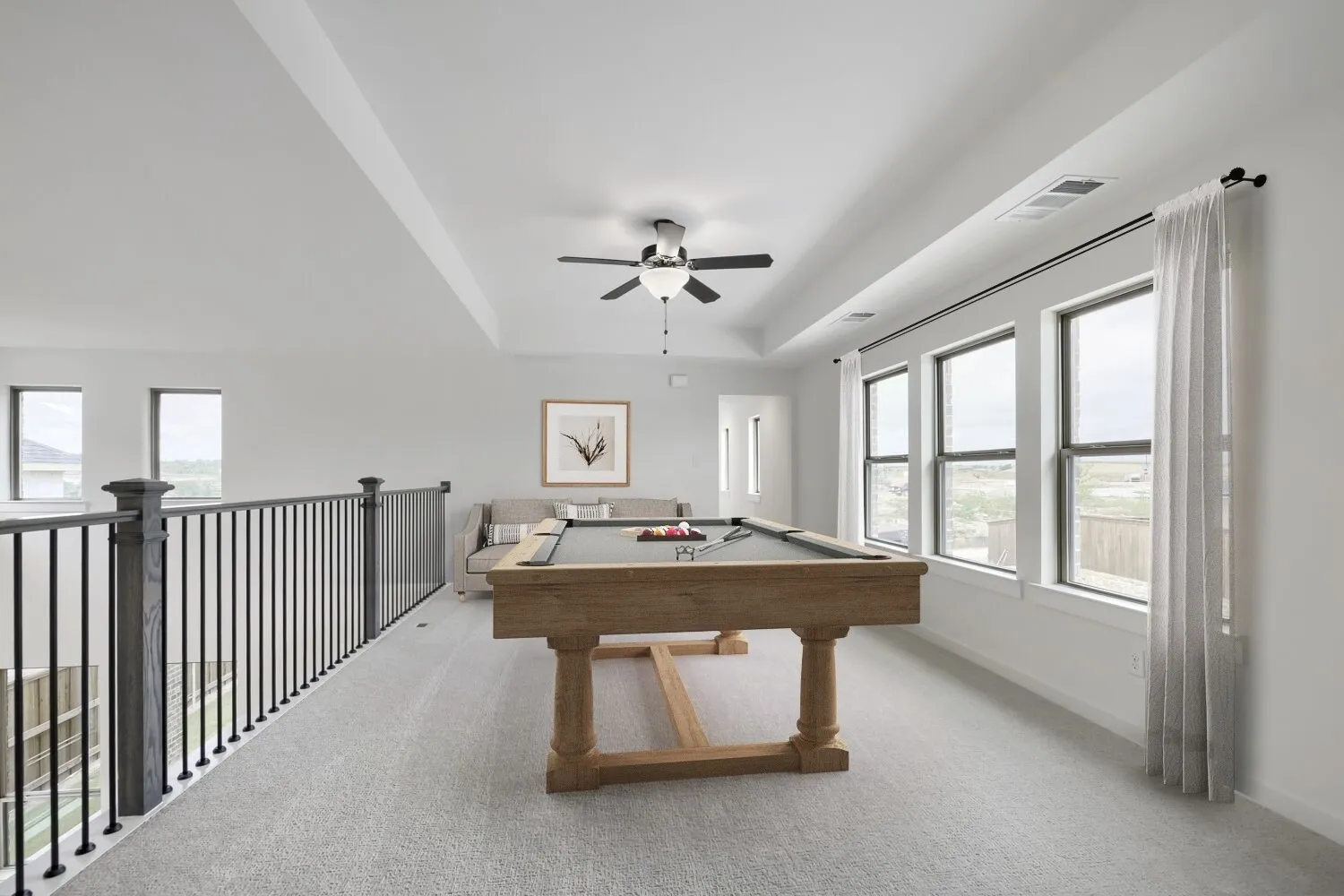 VIRTUALLY STAGED PHOTO Upper-level bonus room featuring carpet flooring, tray ceiling, ceiling fan, multiple windows, and wrought iron railing