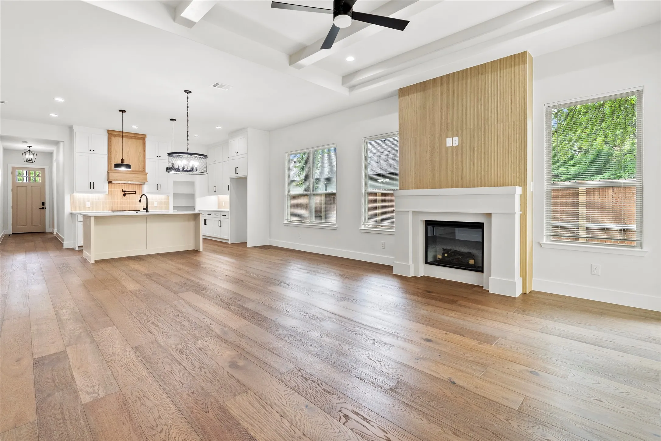 Spacious open-concept interior featuring light wood-finish flooring, a contemporary linear electric fireplace with a white mantel, and a kitchen island with a sink and pendant lighting