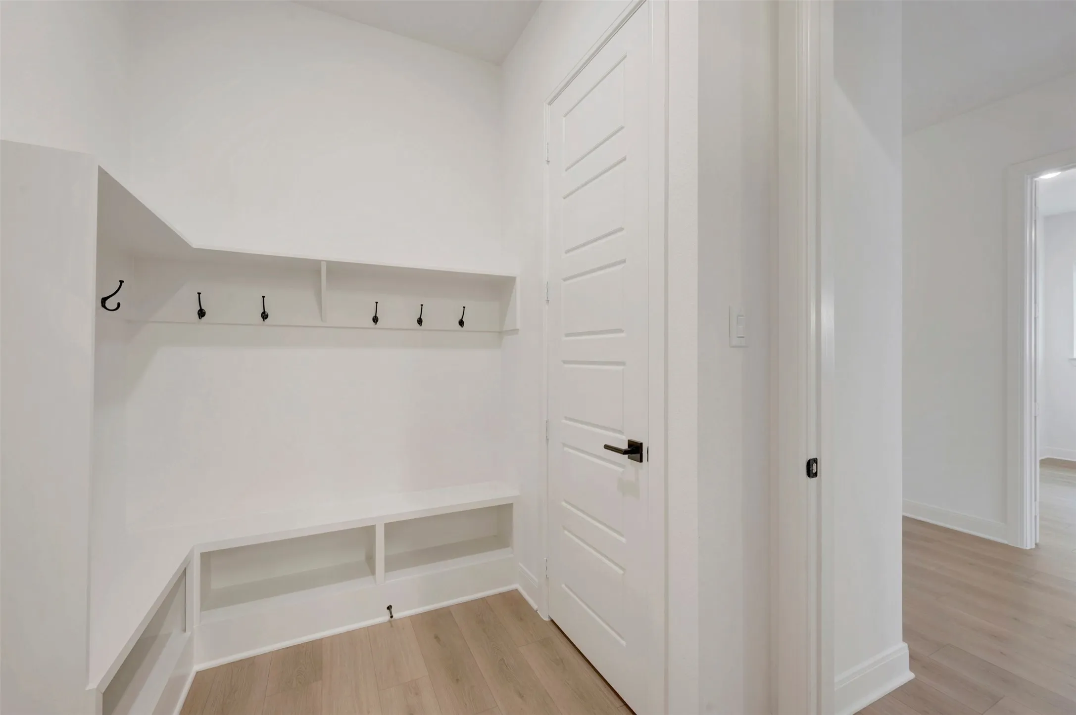 Built-in shelving with hooks, integrated bench seating, wood-finish flooring, white paneled door with matte black hardware, and crisp white walls