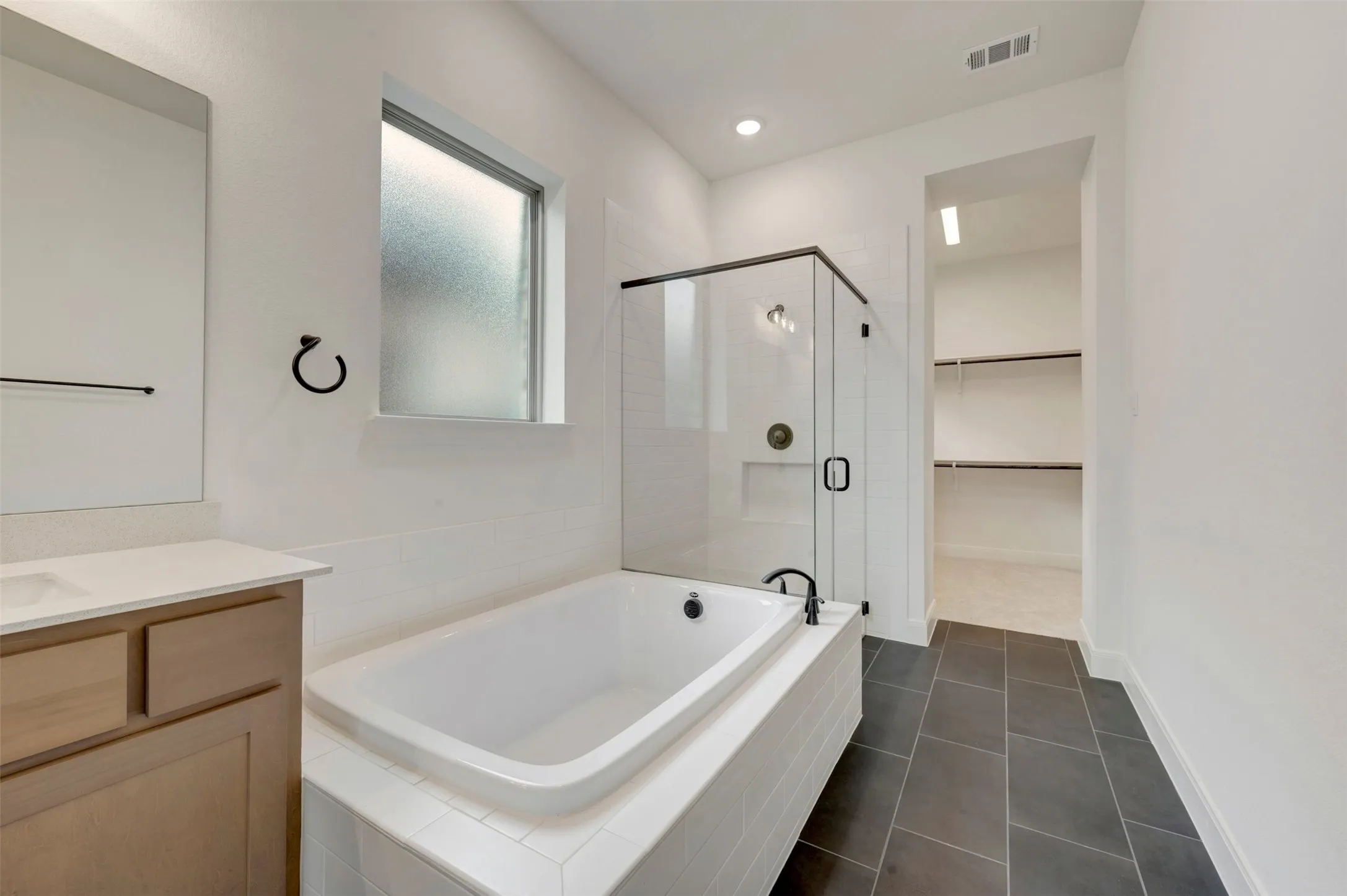 Built-in soaking tub, glass-enclosed shower, single vanity with light-tone cabinetry, dark tile flooring, and recessed lighting