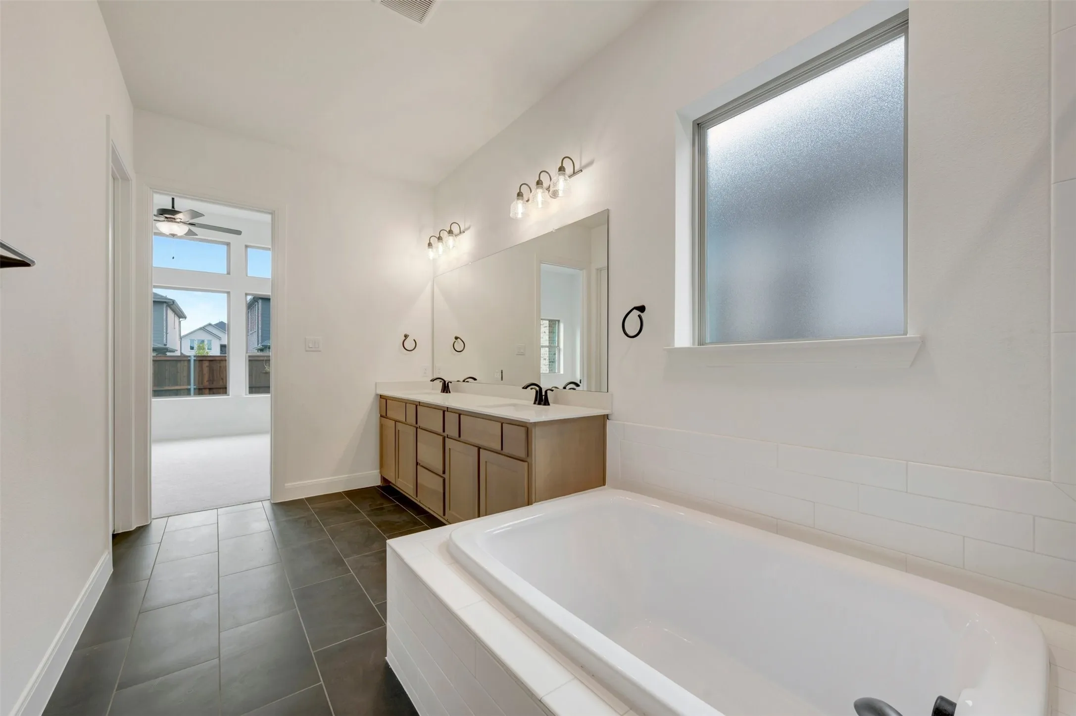 Dual vanity with wood-finish cabinetry, built-in soaking tub, large format tile flooring, and a privacy window