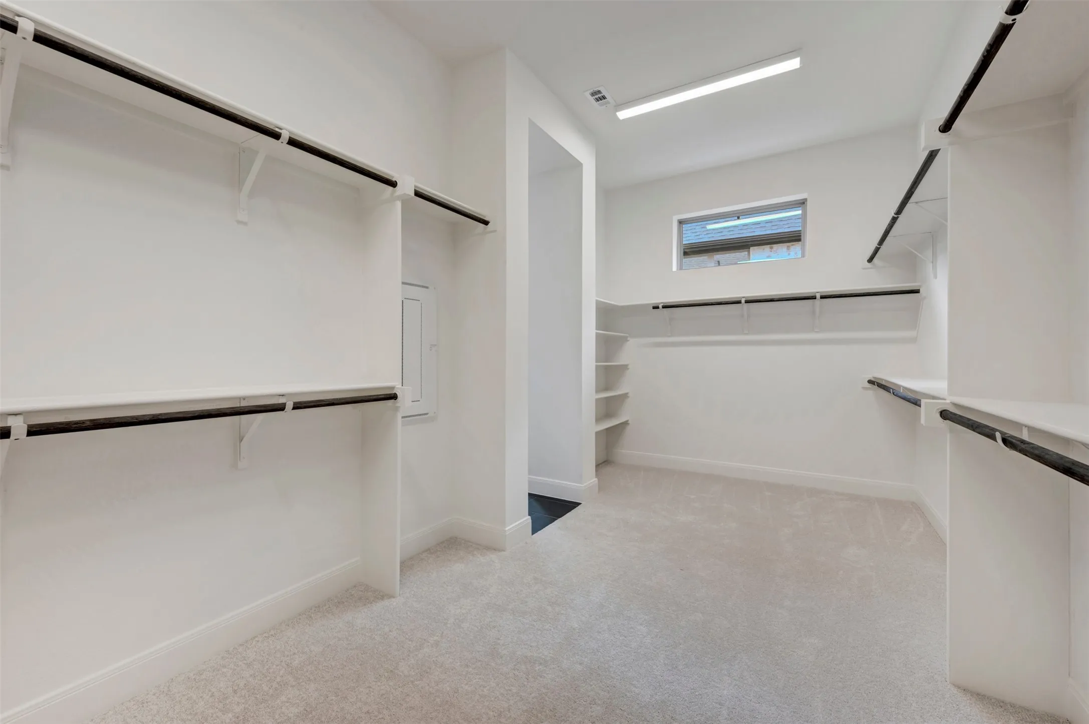 Spacious walk-in closet featuring white built-in shelving, black hanging rods, light-colored carpeting, a high-set window, and recessed lighting