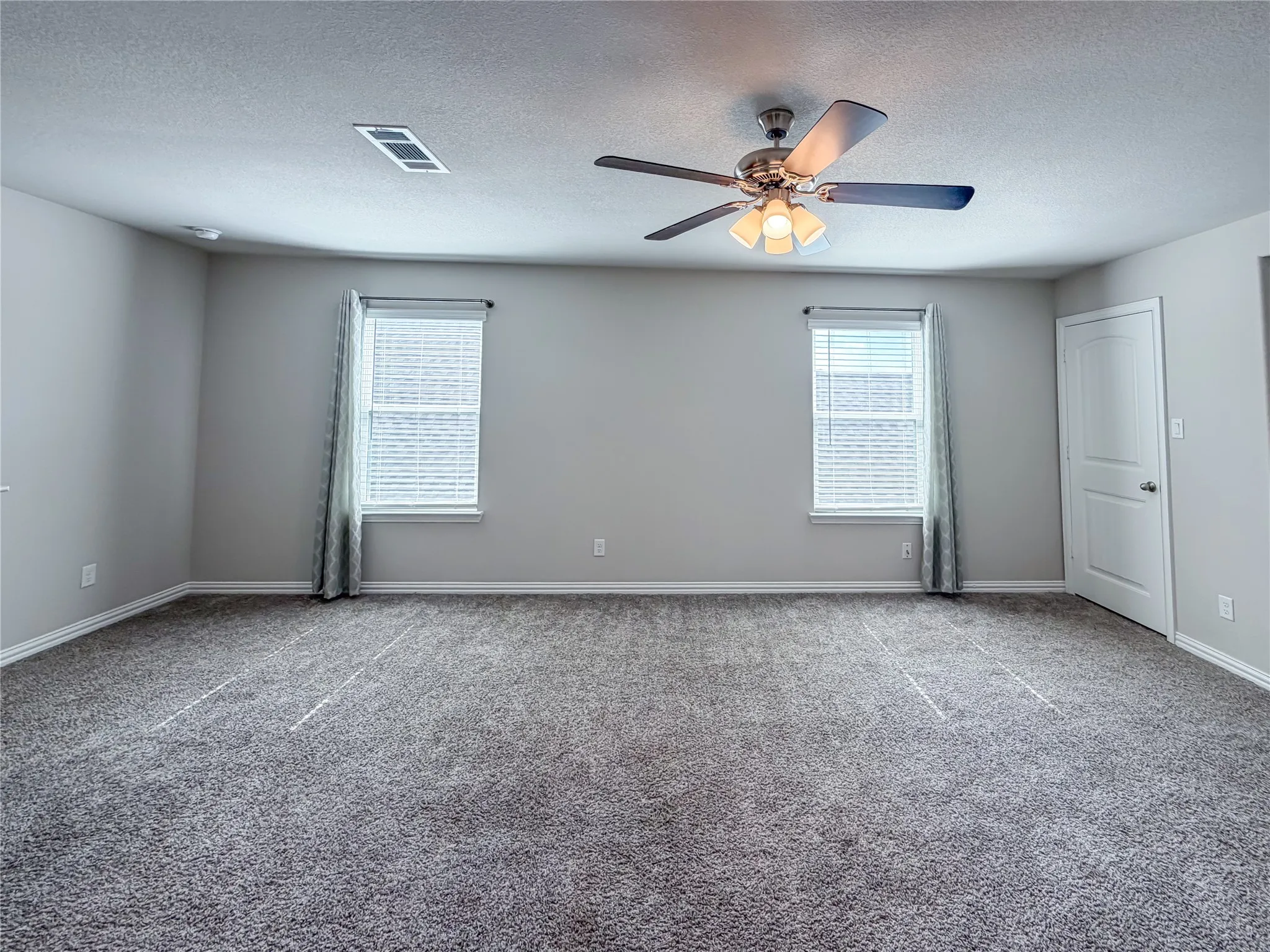 Spacious room featuring gray carpet, light gray walls, two windows with blinds and patterned drapes, a ceiling fan with integrated lighting, and a white paneled door