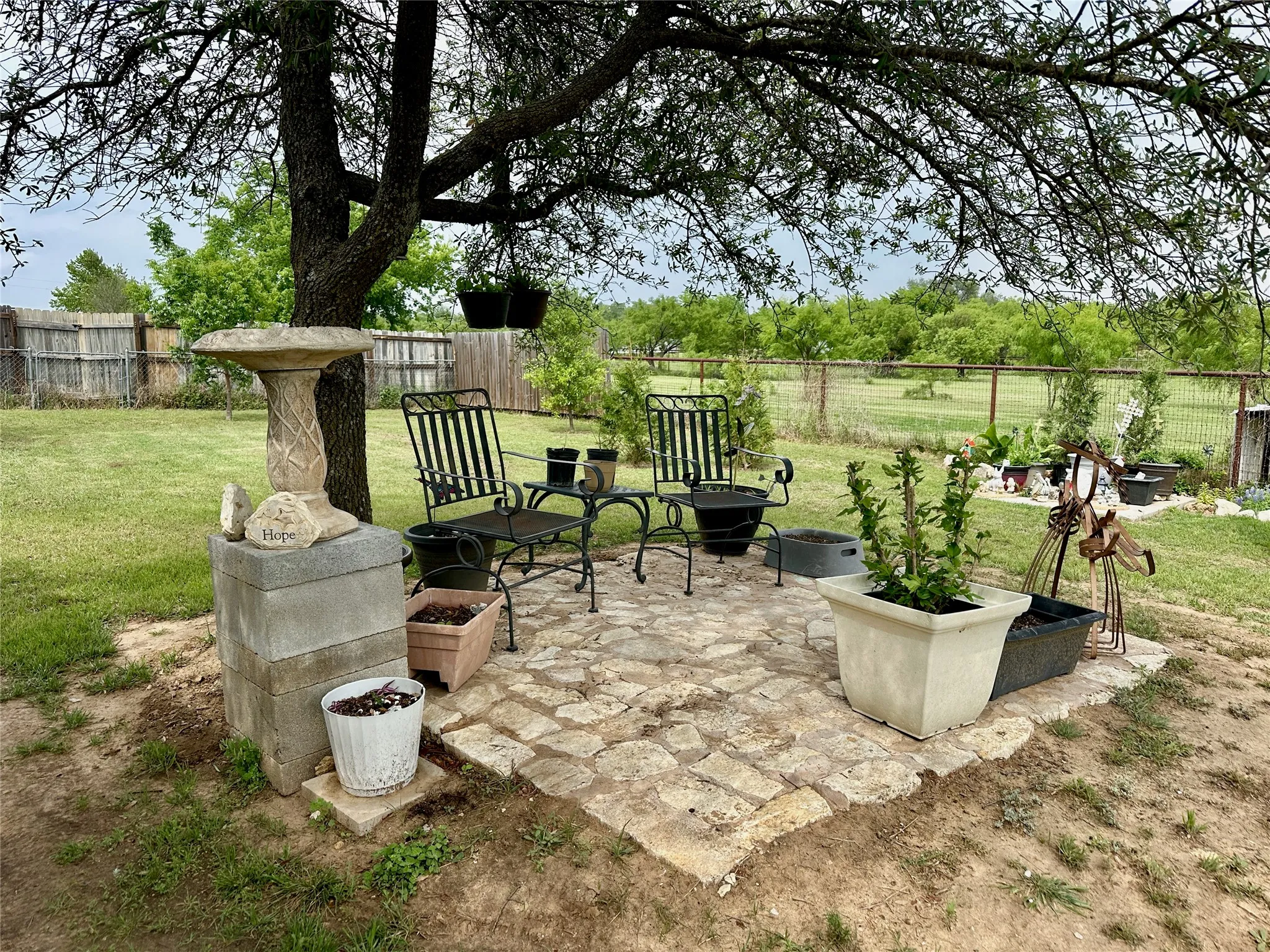 Paved stone patio area beneath a mature tree, featuring a stone bird bath, a metal bistro set, and various plant containers