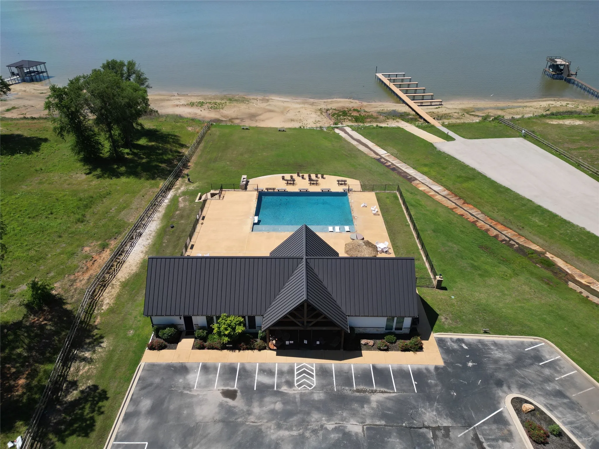 Waterfront building with a dark-colored metal roof, featuring an adjacent swimming pool with a concrete deck, a paved parking area, and direct access to a private dock extending into the water