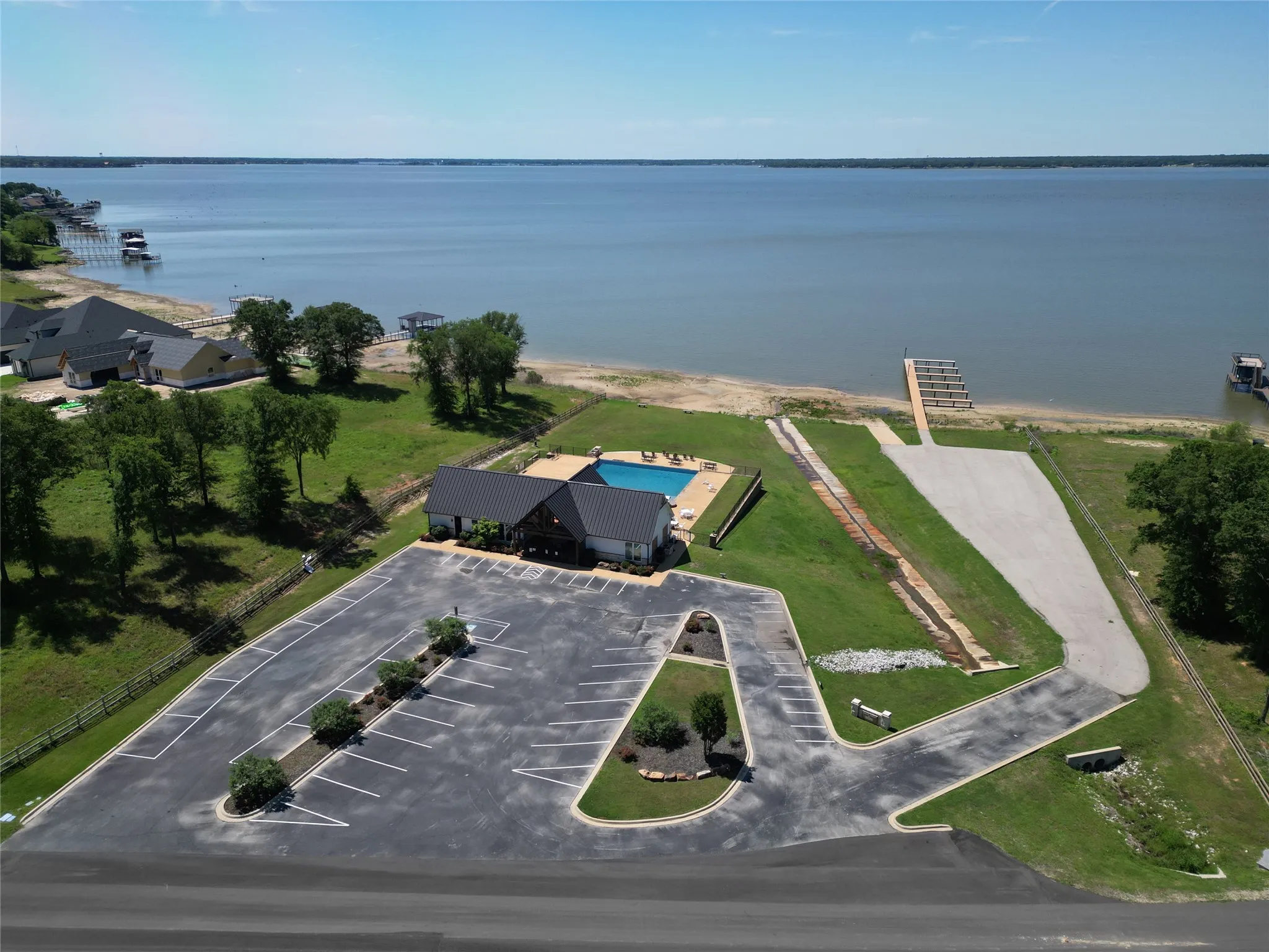 Expansive lakeside property featuring a large parking area, a community building with a dark roof, a swimming pool, a sandy beach, and a boat ramp with a dock