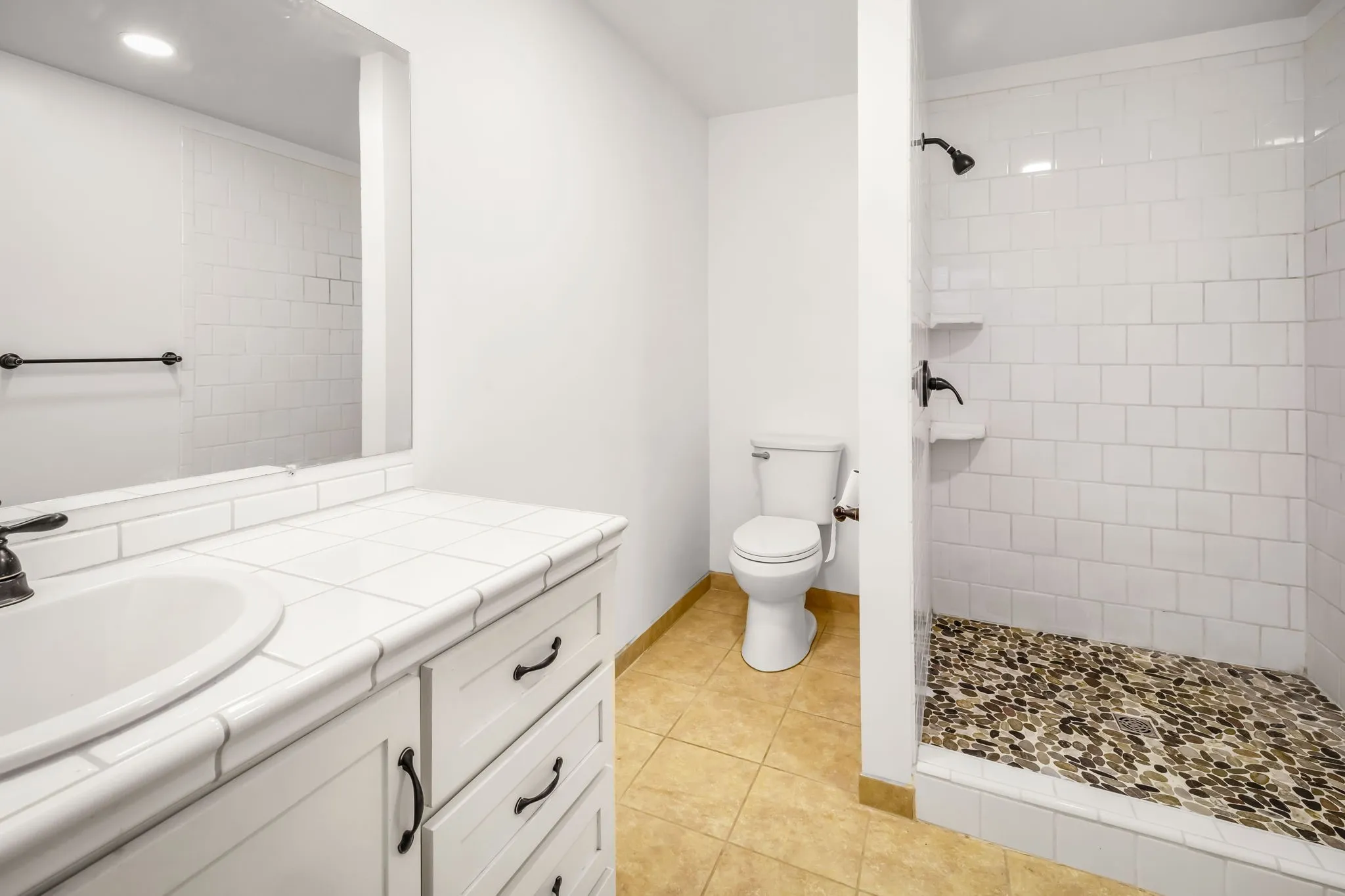 Bathroom featuring a tiled shower with a pebble floor, a white vanity with a drop-in sink, dark bronze fixtures, and warm-toned floor tiles