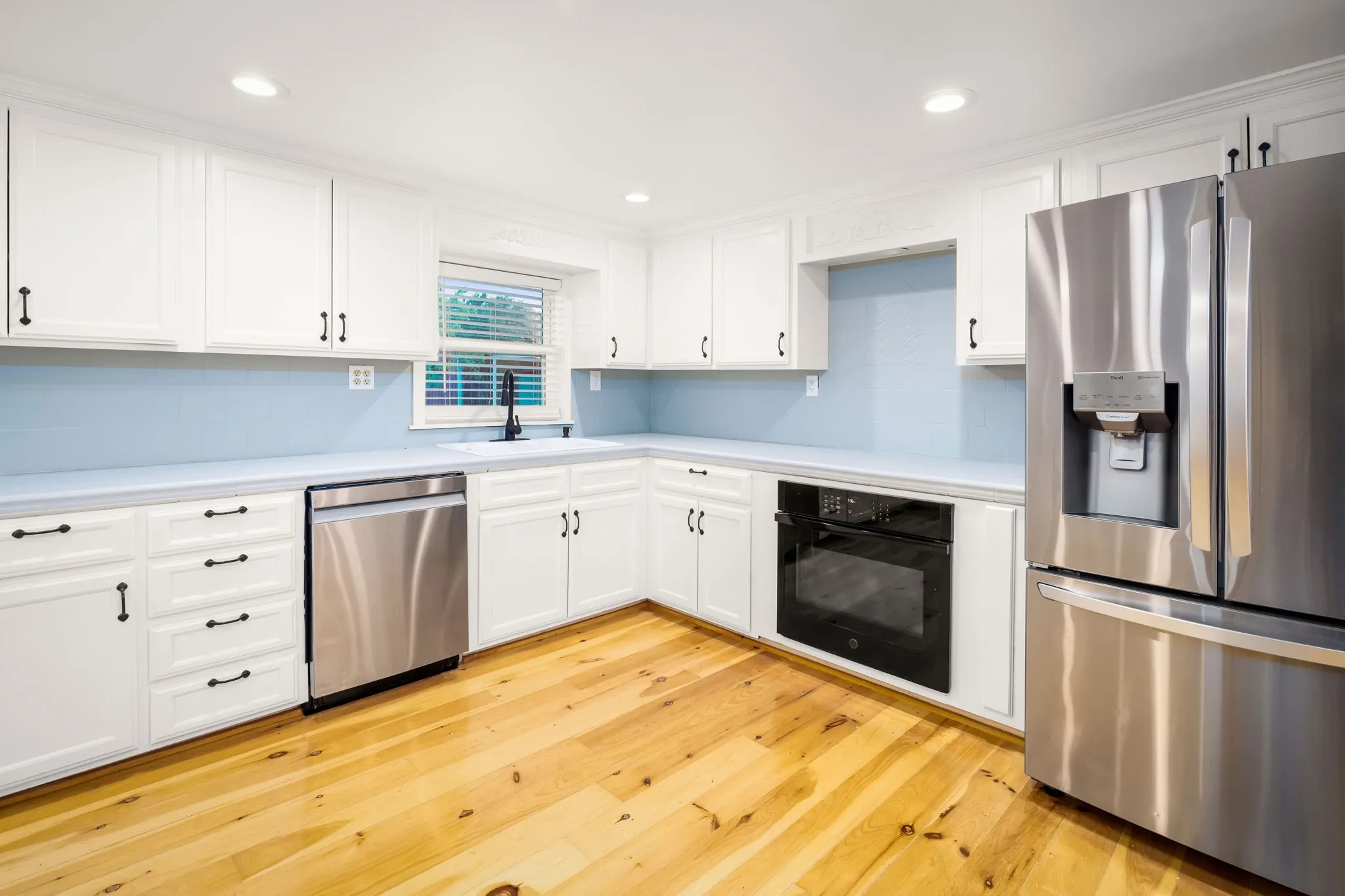 Kitchen featuring wood-finish flooring, white cabinetry with dark hardware, stainless steel appliances, light blue subway tile backsplash, and light-toned countertops