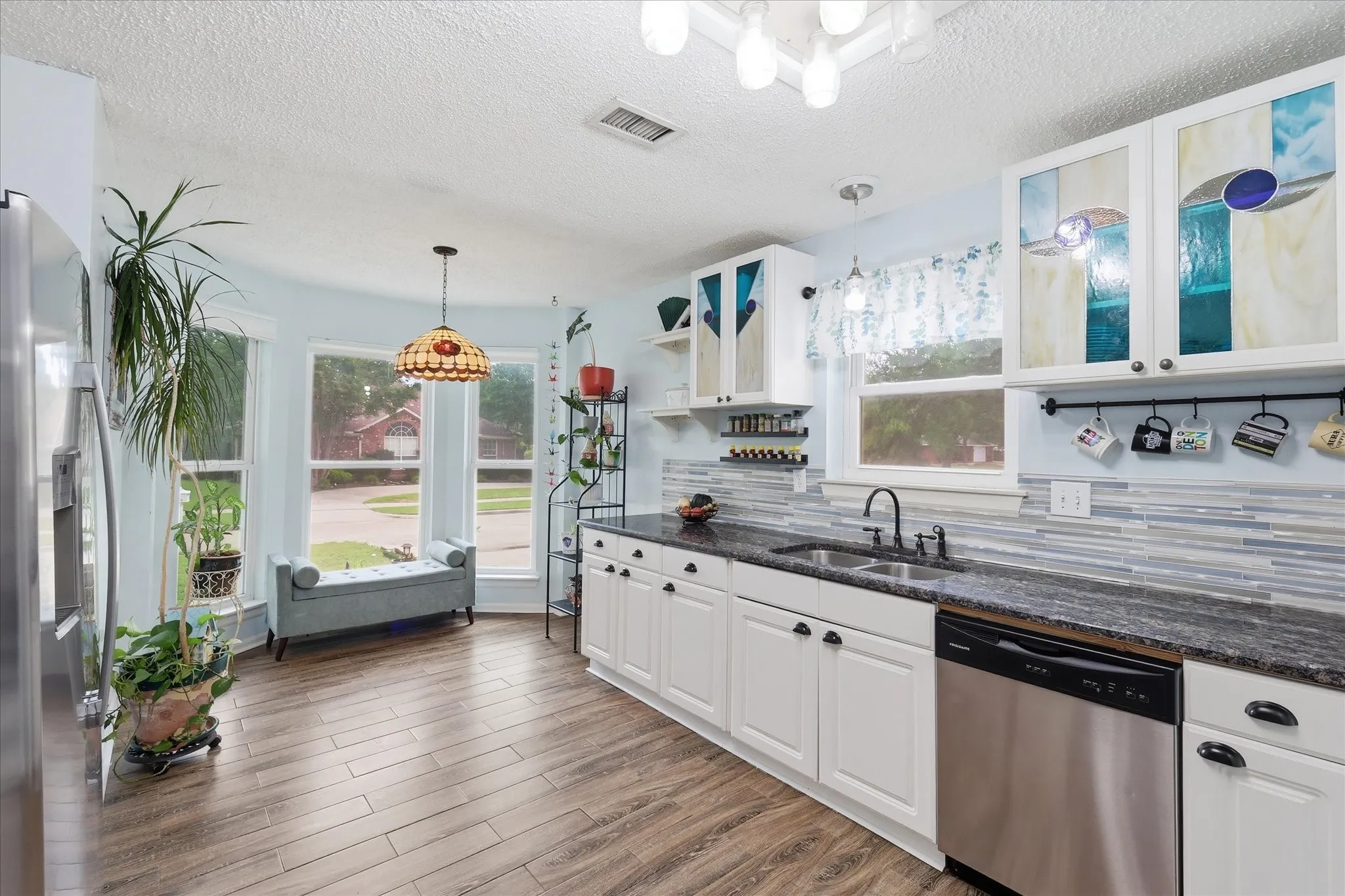 Kitchen featuring wood-finish flooring, white cabinetry with decorative glass inserts, dark countertops, and a tiled backsplash