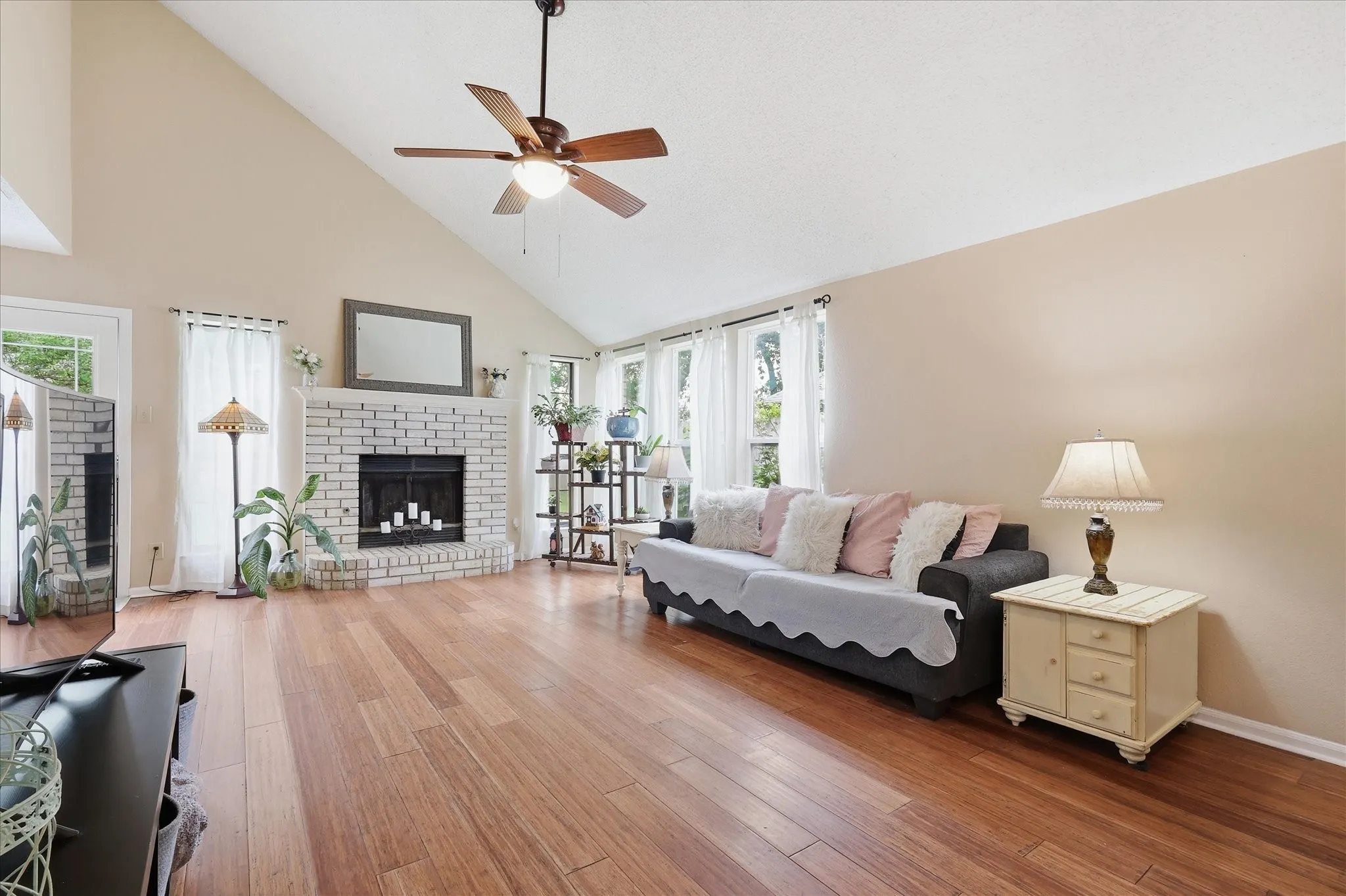 Spacious living area featuring high vaulted ceilings, warm wood-finish flooring, a white brick fireplace, multiple windows with natural light, and a ceiling fan
