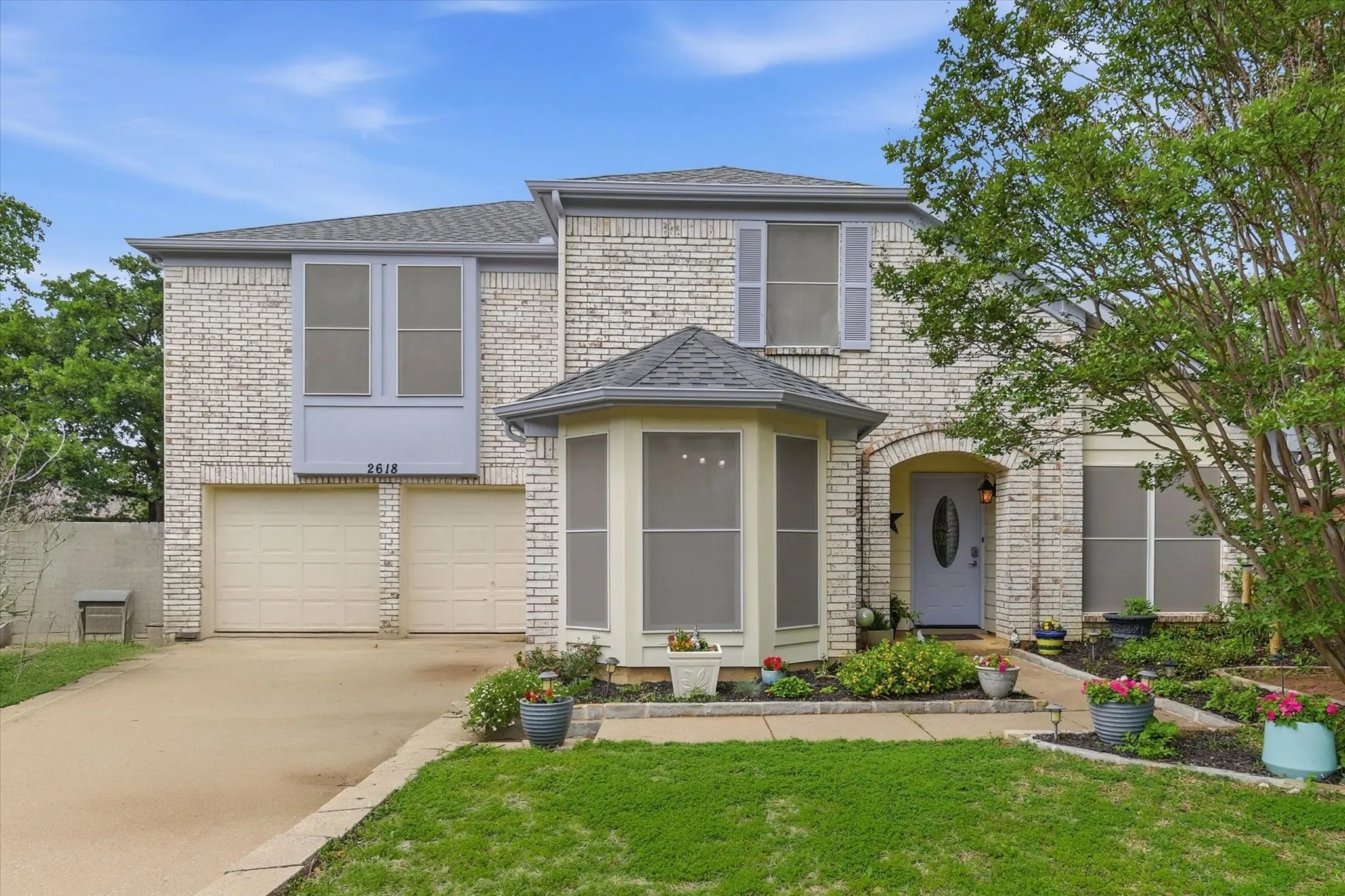 Two-story brick facade residence featuring a two-car garage, a bay window with a shingled roof, an arched entry, and a light blue front door with an oval glass insert