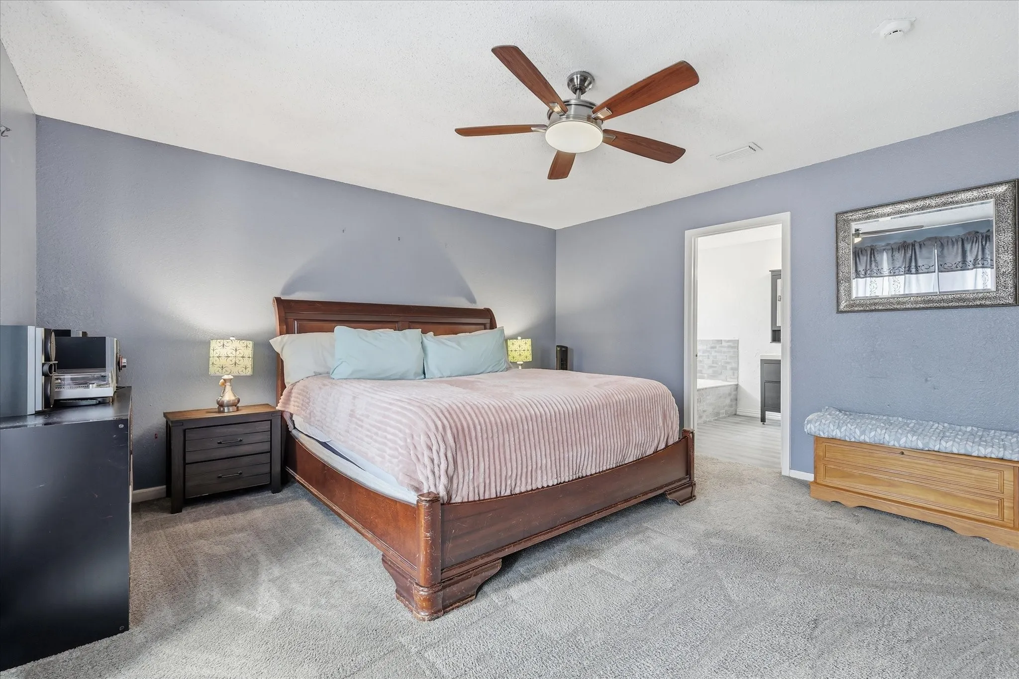 Carpeted bedroom featuring a ceiling fan with integrated lighting, a spacious bed frame, and an open doorway revealing a tiled bathroom with a built-in tub