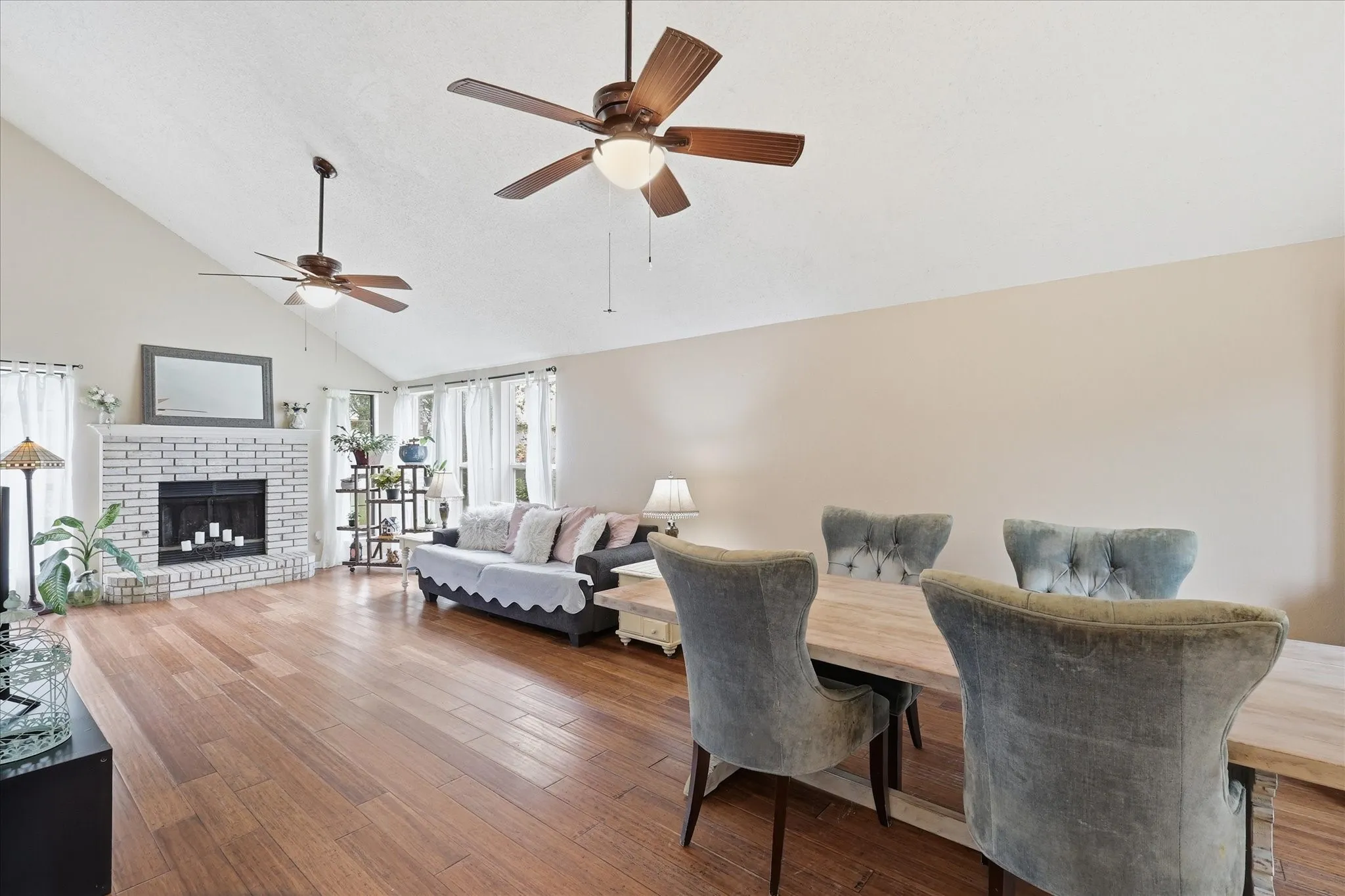 Open-concept living area featuring vaulted ceilings, wood-finish flooring, a white-painted brick fireplace, and multiple windows