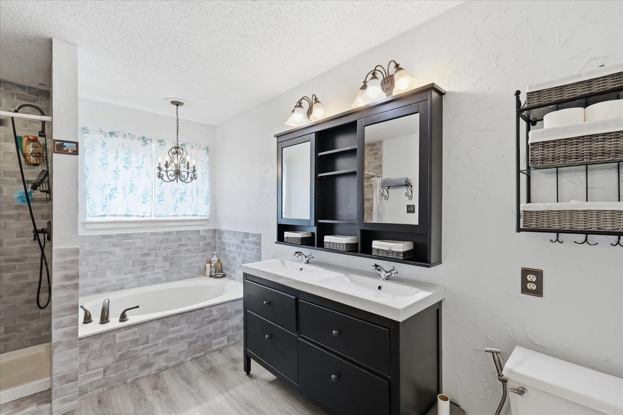 Bathroom featuring a double vanity with integrated sinks, a built-in tub with tile surround, a separate shower with gray tile, wood-finish flooring, and a decorative chandelier