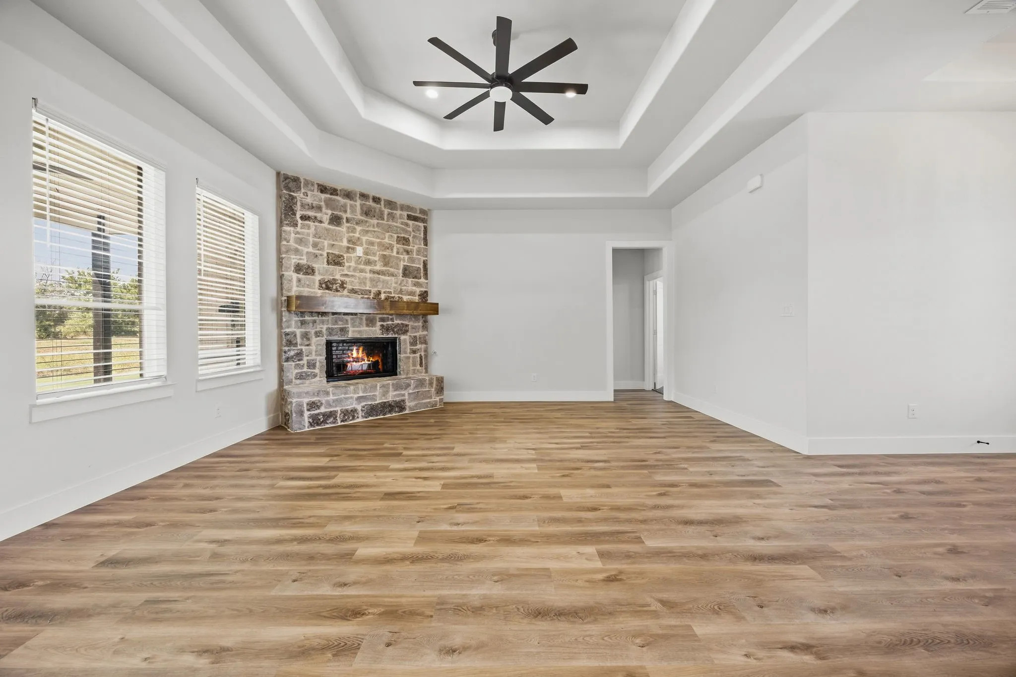 Tray ceiling with a ceiling fan, stone-clad fireplace with a wood mantel, wood-finish flooring, and multiple windows