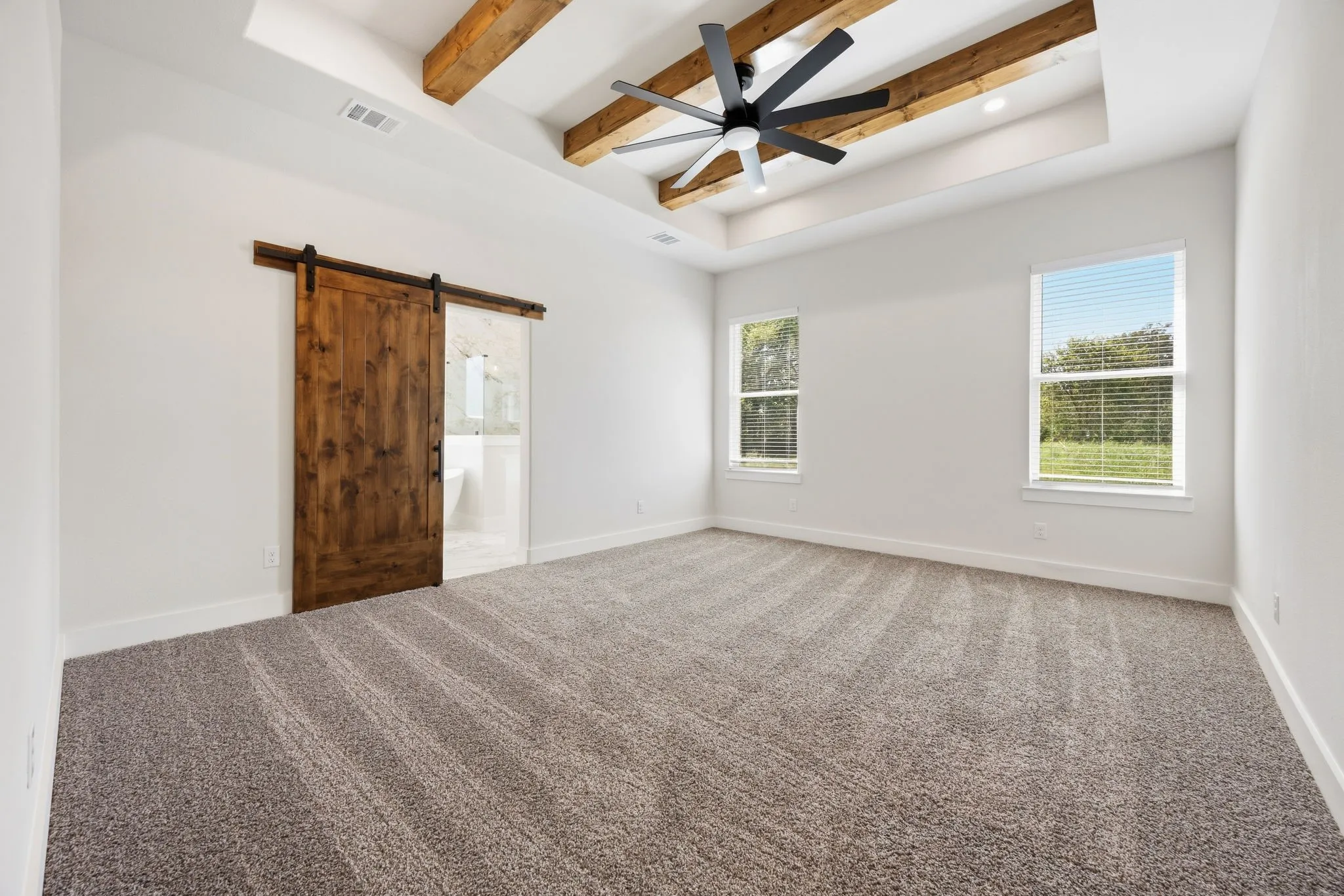 Tray ceiling with exposed wood beams, modern ceiling fan, wood-finish barn door, and two windows with blinds