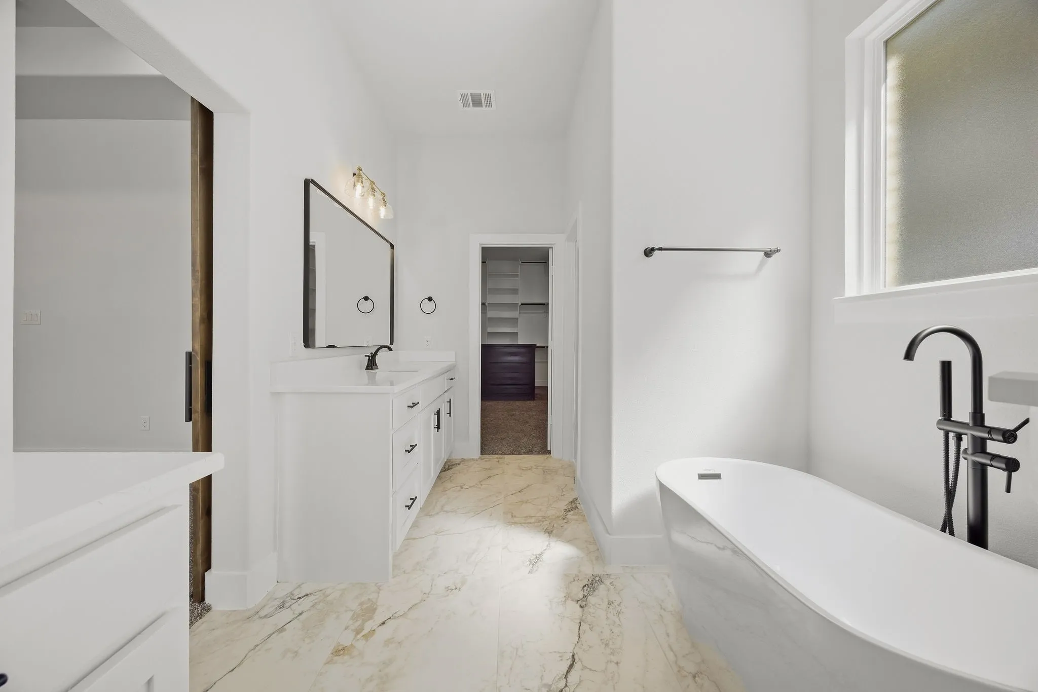 White vanity with integrated sink, black framed mirror, freestanding tub with matte black fixtures, and patterned tile flooring