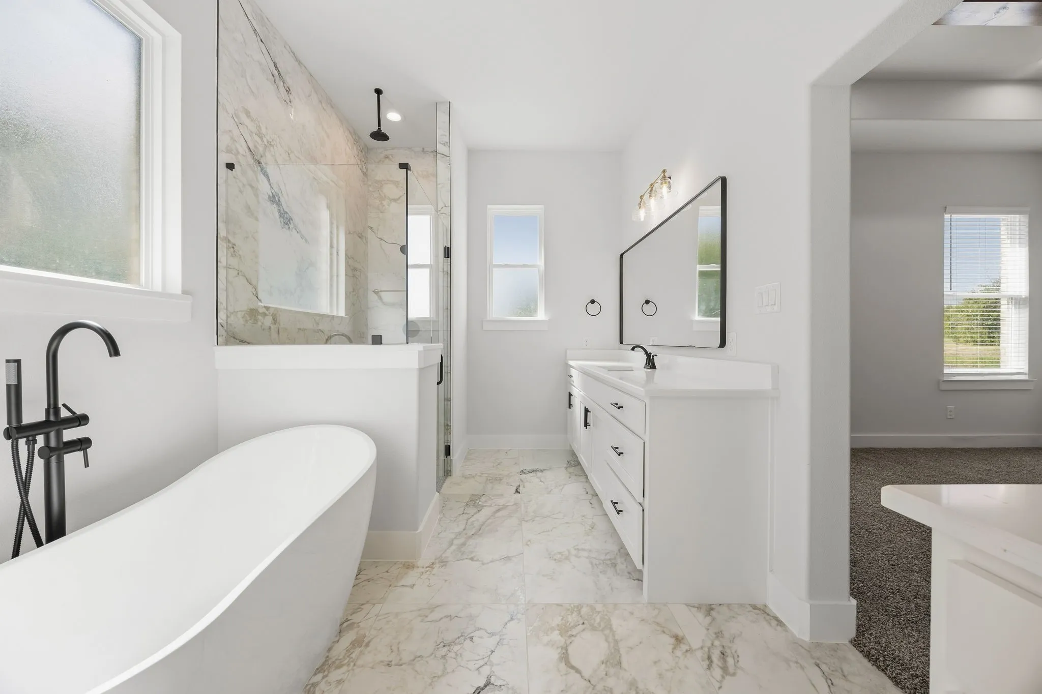 Freestanding soaking tub with a floor-mounted black faucet, glass-enclosed shower with a rain shower head, dual-sink vanity with white cabinetry, and marble-finish tile flooring