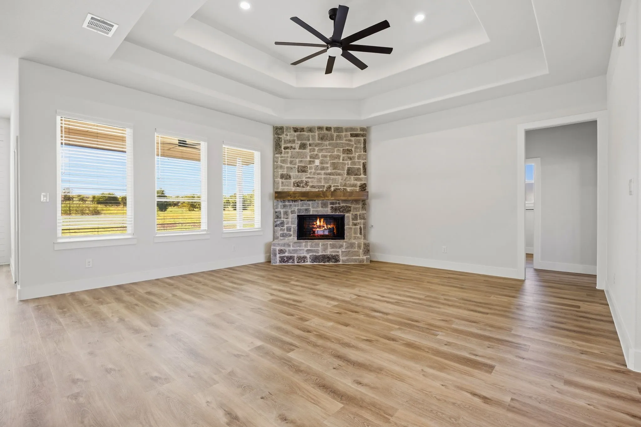 Tray ceiling with recessed lighting and a ceiling fan, stone-clad corner fireplace with wood mantel, three large windows, and wood-finish flooring