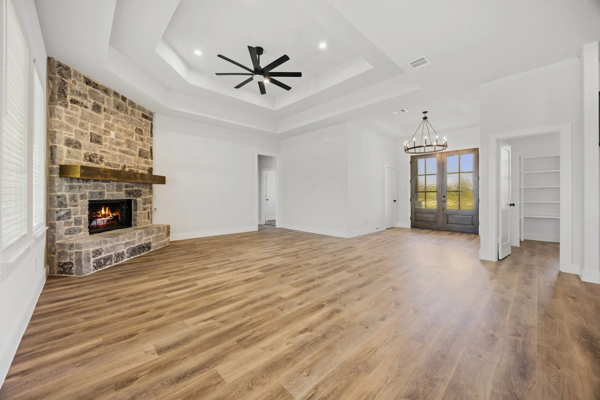 Spacious room featuring wood-finish flooring, a stone fireplace with a wood mantle, a tray ceiling with a ceiling fan, and double entry doors with a transom window