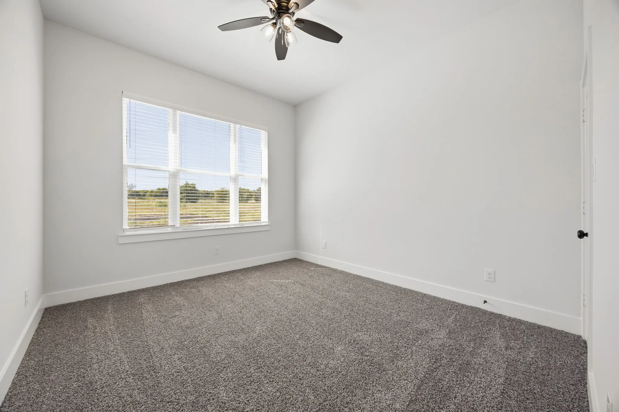 Spacious room featuring neutral carpeting, white walls, a large multi-pane window with blinds, and a ceiling fan