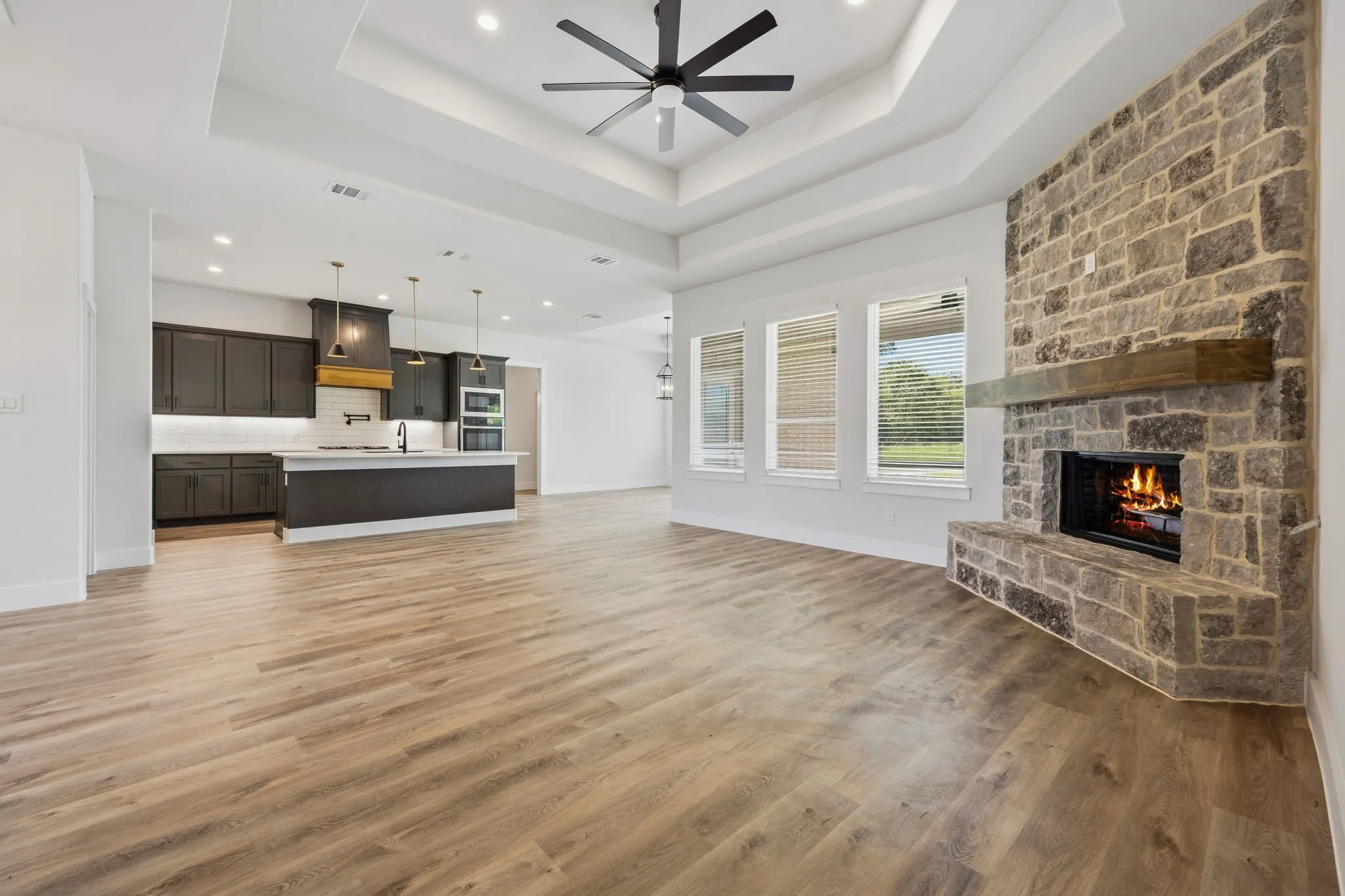Open-concept living space featuring wood-finish flooring, a stone-faced fireplace with a wood mantle, a tray ceiling with a ceiling fan, and a kitchen with dark cabinetry