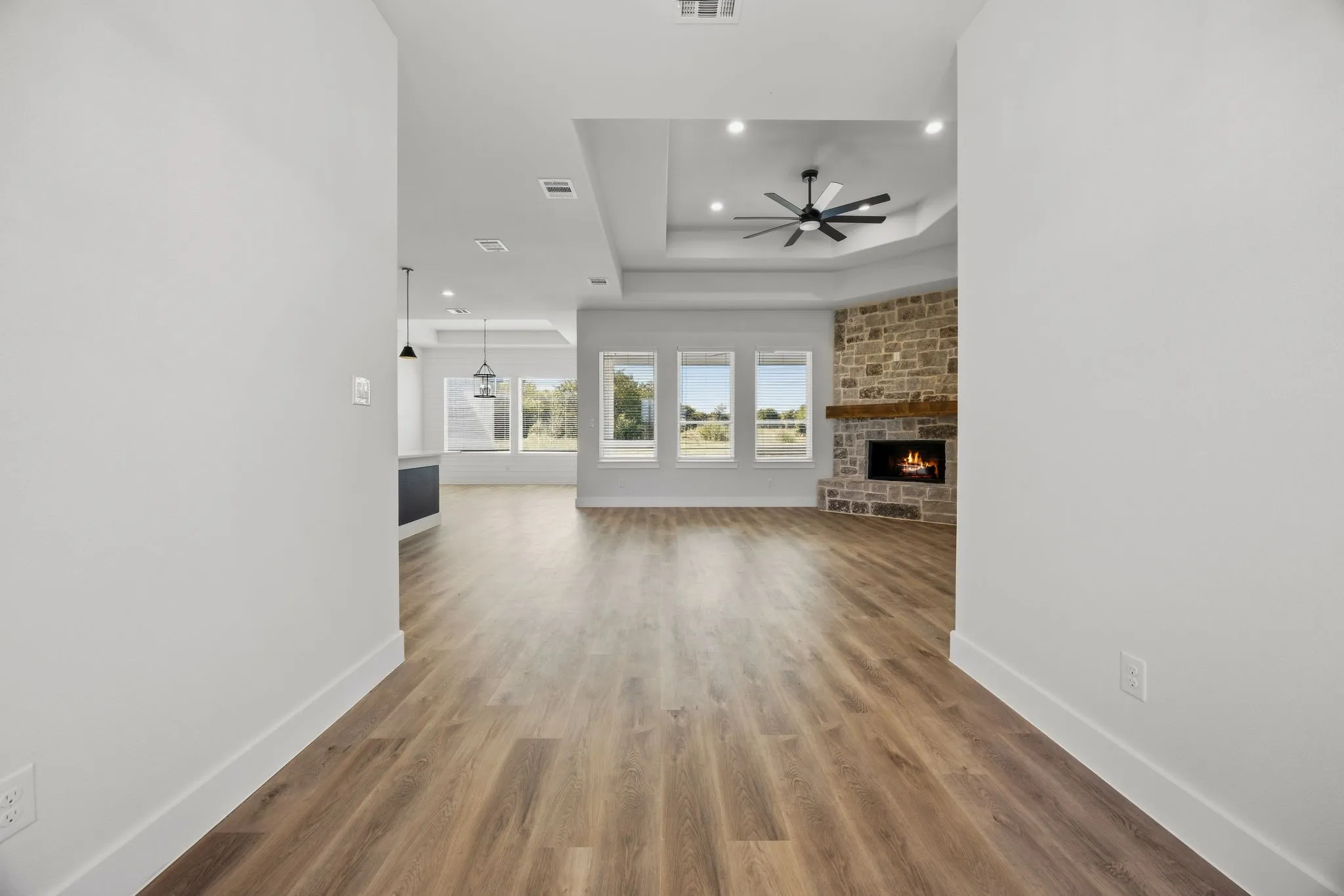 Expansive open-concept living space with wood-finish flooring, a stone-faced fireplace with a wood mantel, a tray ceiling with a ceiling fan, and multiple windows