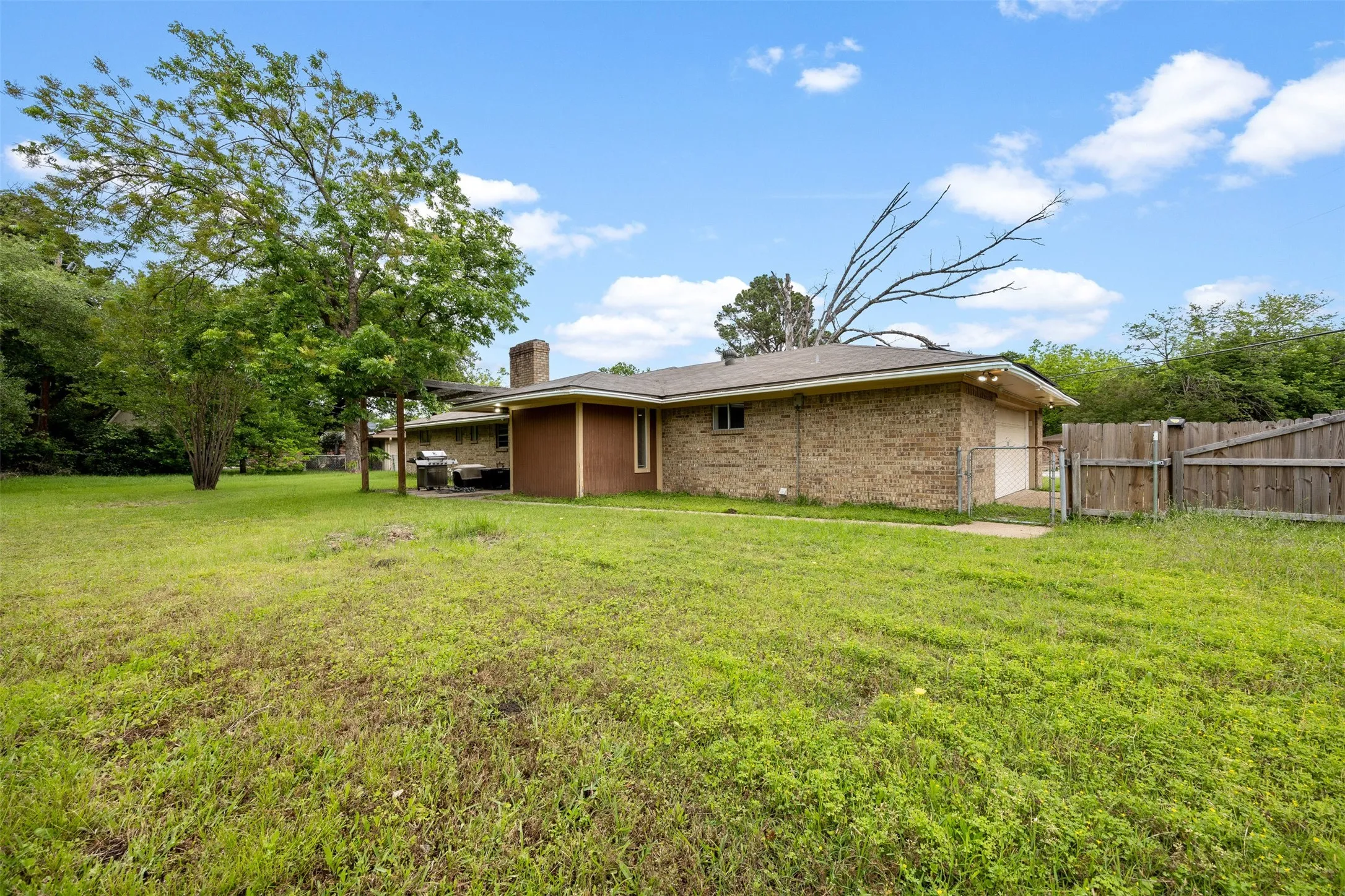 Expansive backyard featuring a large brick exterior home, mature trees, a wooden privacy fence, and an attached covered patio