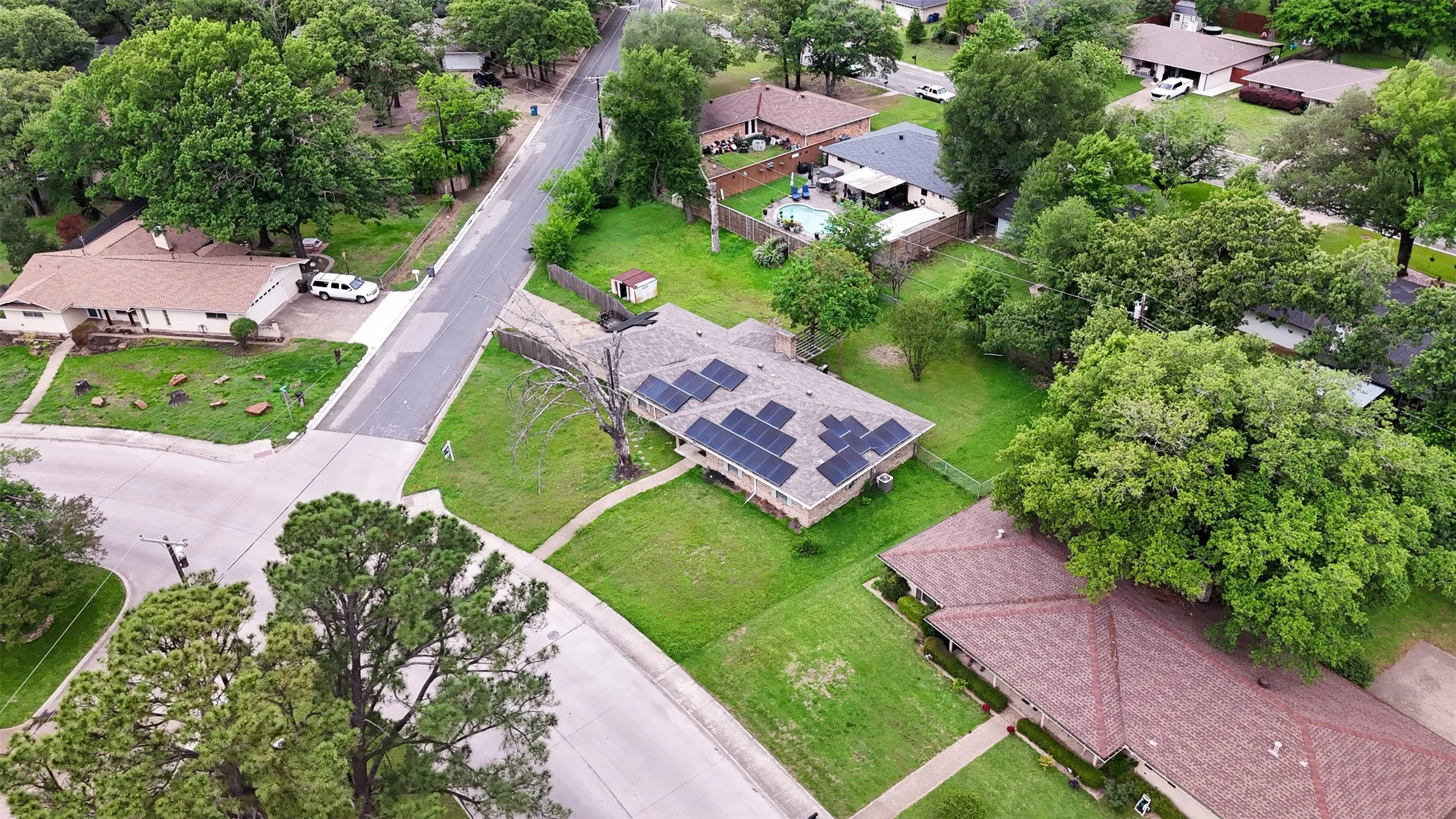 Ranch-style residence featuring a brown shingle roof with solar panel installation, a green lawn, and mature trees