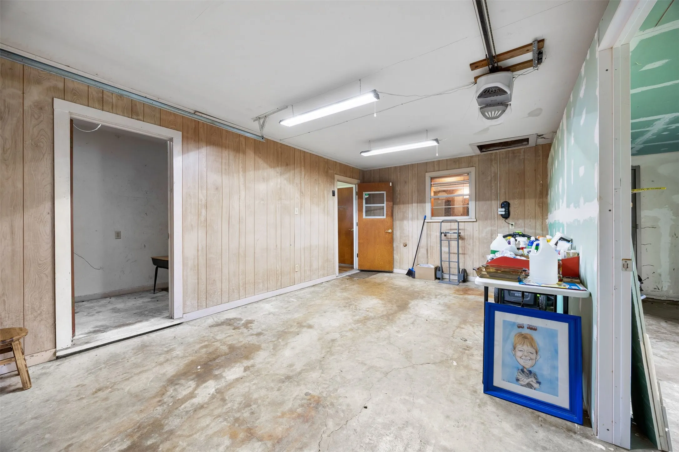 Spacious utility area featuring wood-paneled walls, concrete flooring, fluorescent overhead lighting, and a visible garage door opener mechanism