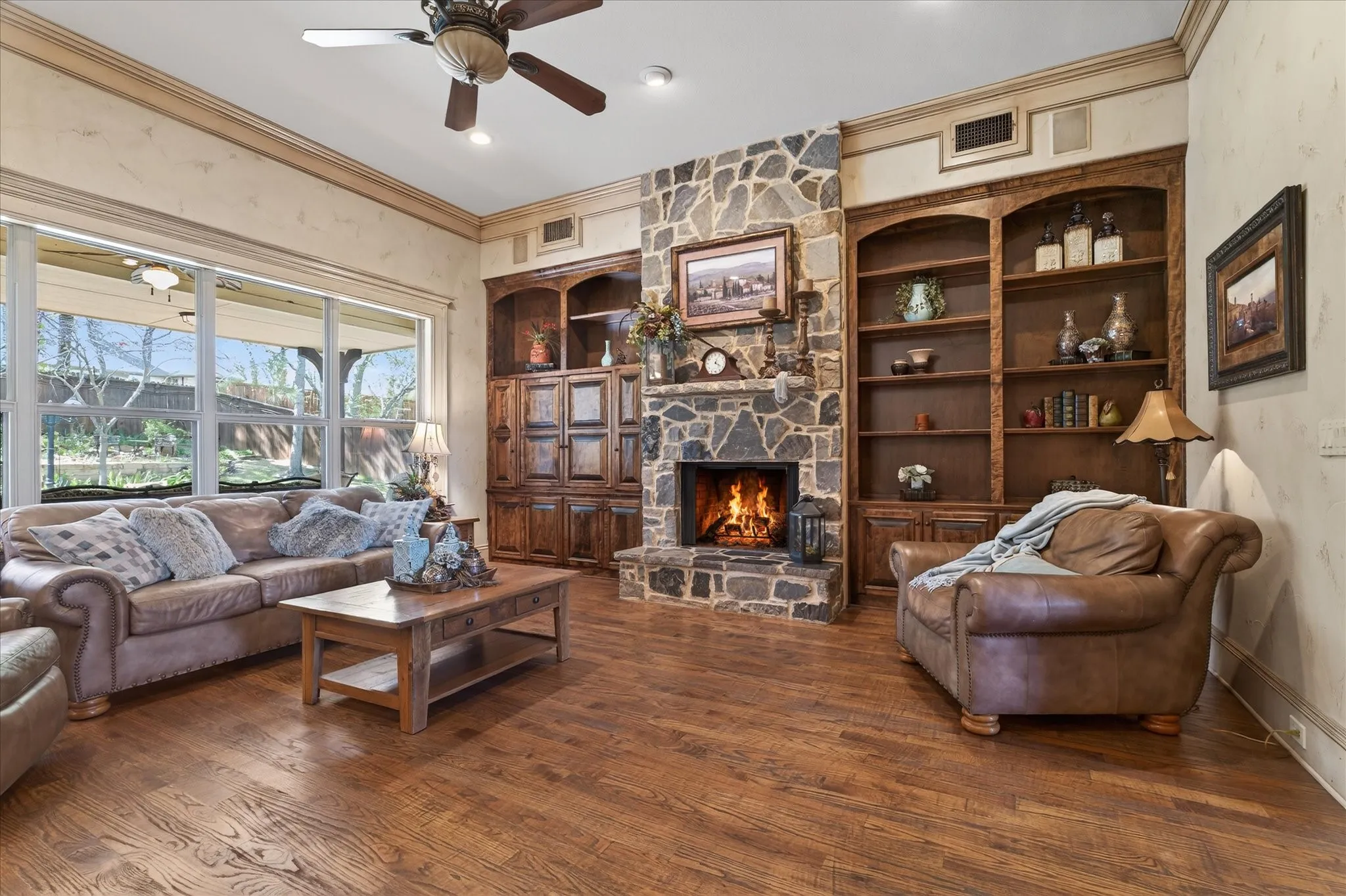 Spacious living area featuring a floor-to-ceiling stone fireplace, custom built-in cabinetry, wide-plank wood-finish flooring, and a multi-pane picture window