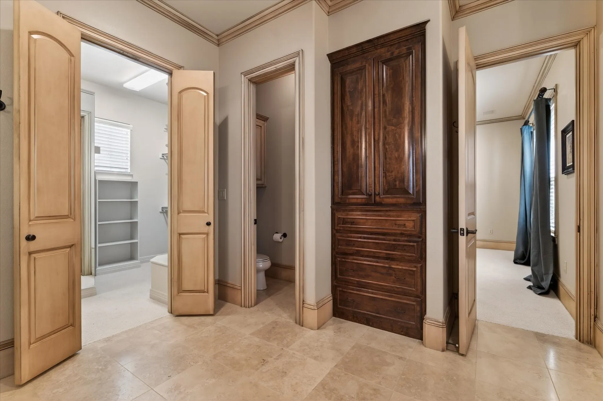 Travertine flooring throughout a hallway featuring an integrated dark wood-finish storage cabinet, a bathroom with an open shower and built-in shelving, and a separate powder room