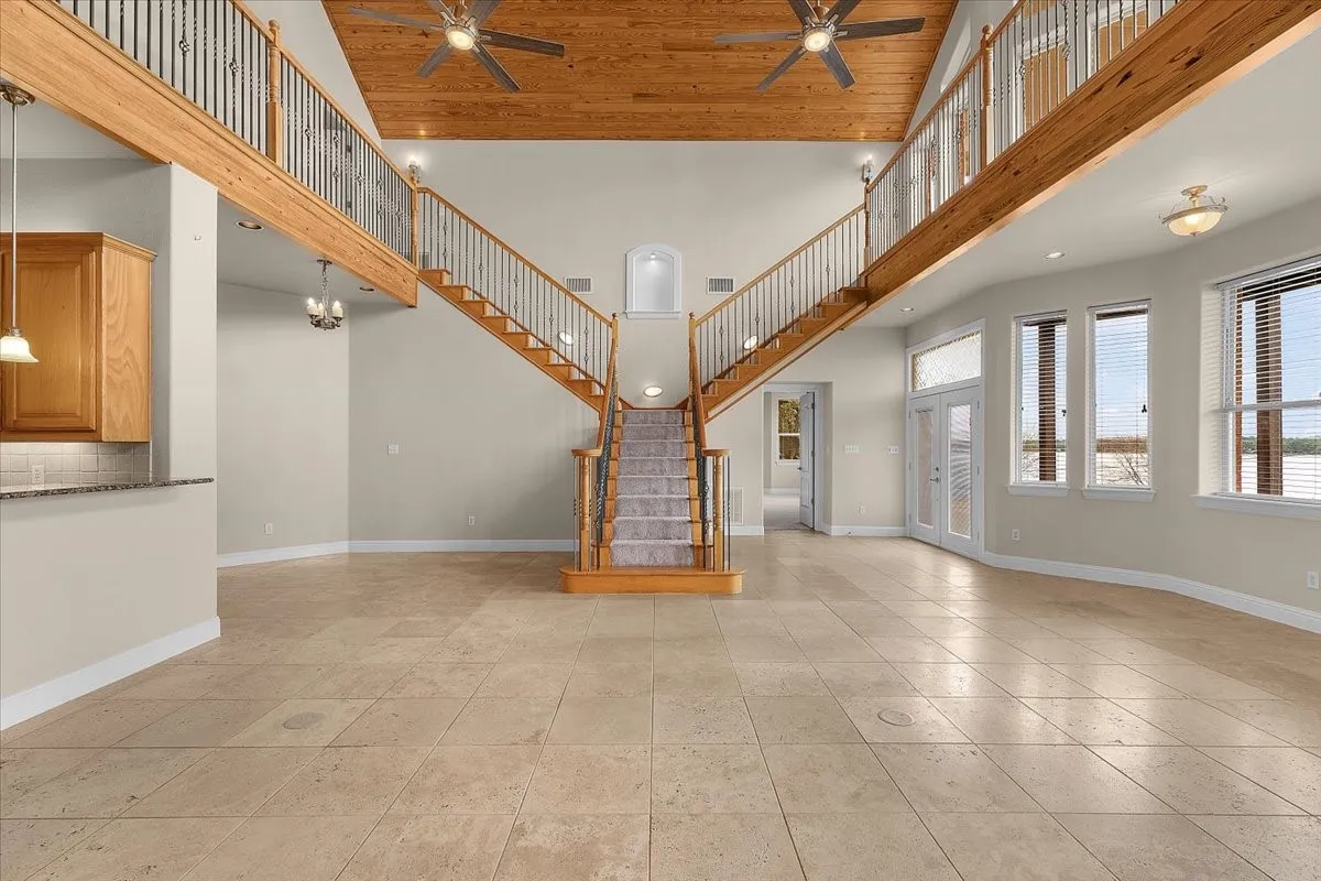 Spacious living area featuring a bifurcated staircase with carpeted treads and wrought iron balusters, a wood-paneled vaulted ceiling with ceiling fans, large windows with blinds, a built-in bar counter with wood cabinetry and tile backsplash, and light-toned floor tiling