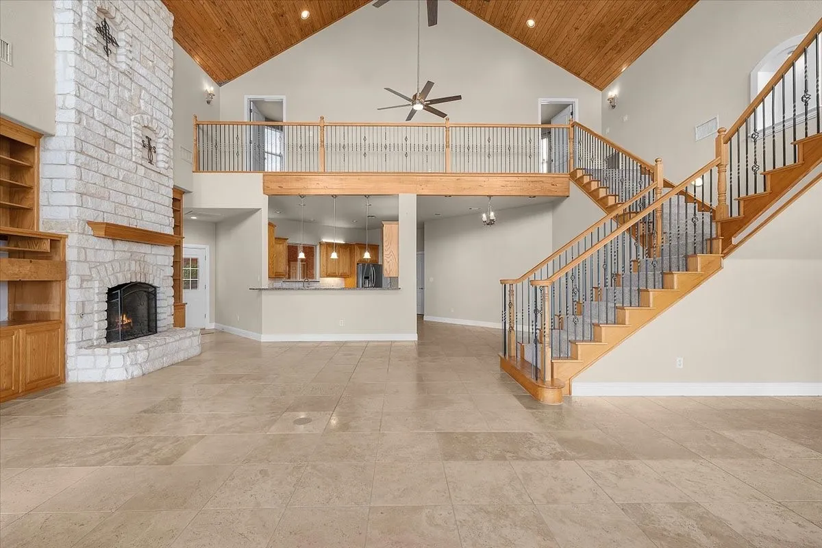 Vaulted ceiling with wood-finish planks, dual-sided staircase with wrought iron balusters, stone fireplace with a wood mantle, built-in wooden shelving, and large format tile flooring